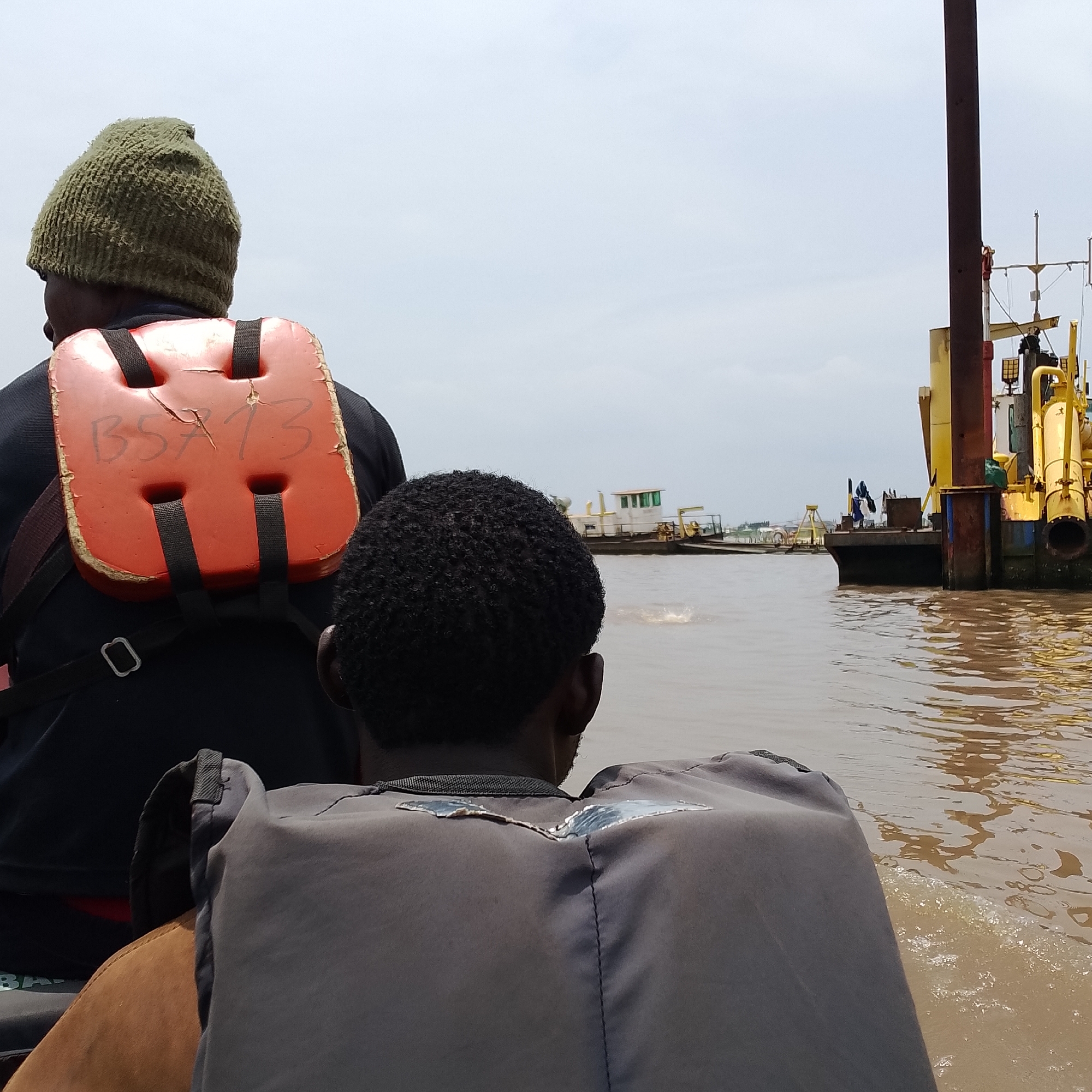 Two individuals, viewed from behind and from the back of a vessel, are on a body of water, possibly a river. The person in the foreground is wearing a grey life vest with a metallic emblem or patch on the back. The person in the background is wearing a green knitted cap and an orange life vest with "BSA 73" written on it in black marker. In the background, there are several boats and barges, including a large yellow barge with machinery, a smaller white and green boat, and other vessels. The water is muddy brown, suggesting it is a river or estuary. The sky is overcast. The scene is consistent with a riverine environment in a populated area, such as the waters near Ado, Nigeria, where industrial barges and local boats are common.