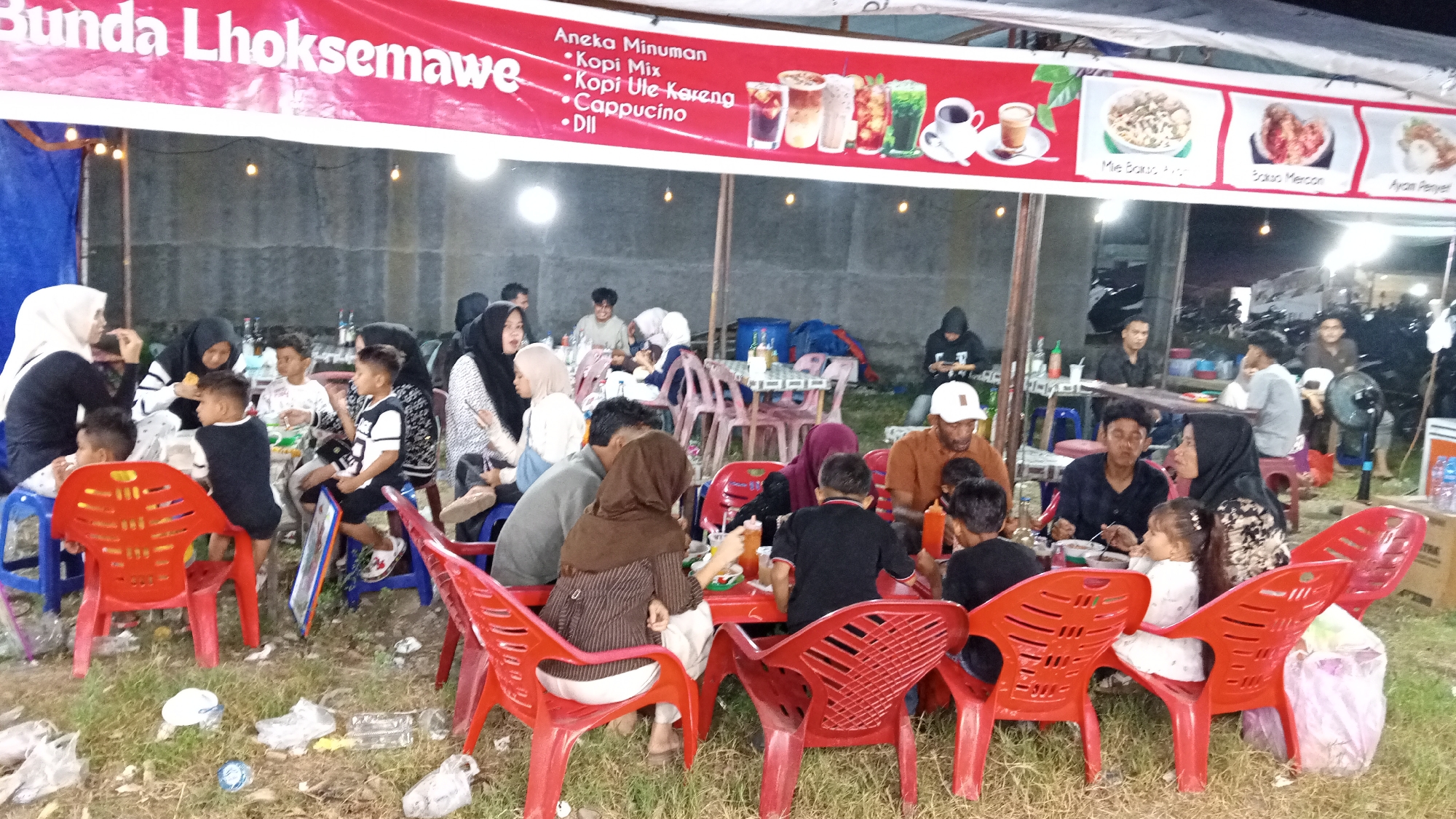 The image depicts an outdoor food stall or eatery at night, likely in Blangcotbaroh, Indonesia, as indicated by the text on the banner which reads "Bunda Lhokse-mawe". The banner features illustrations of various drinks and food items. The foreground is dominated by red plastic chairs and tables, some occupied by people. In the center of the image, a family, including adults and children, is seated around a table, appearing to be eating. Several individuals, dressed in casual clothing and headscarves, are visible at various tables. The background shows more seating areas, people, and parked motorcycles, suggesting a busy evening at a local establishment. The ground is covered with grass and scattered litter.