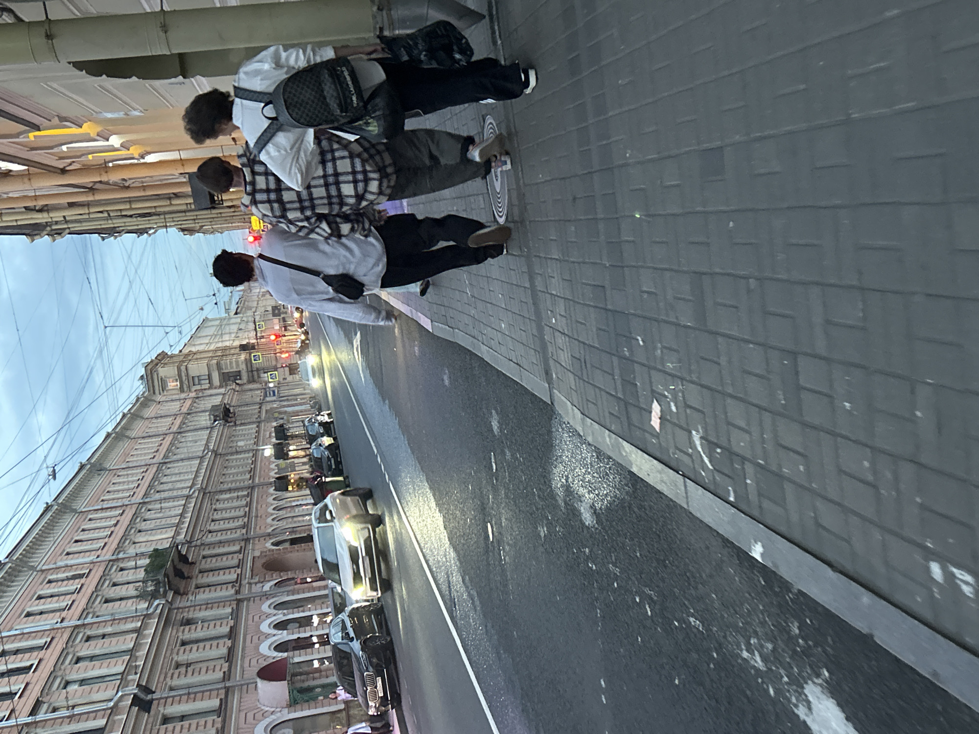 Three young men walk away from the camera on a city sidewalk alongside a street with traffic. The man on the left wears a light-colored shirt and a dark crossbody bag. The man in the middle wears a plaid shirt and dark pants. The man on the right wears a white shirt and a black backpack with a checkered pattern. They are walking in the same direction, suggesting they are together. The street is wet, possibly from recent rain, and cars are driving on it. Buildings line the street on the left side, with shopfronts and windows visible. The sky is overcast and shows signs of twilight. The sidewalk is paved with gray bricks.