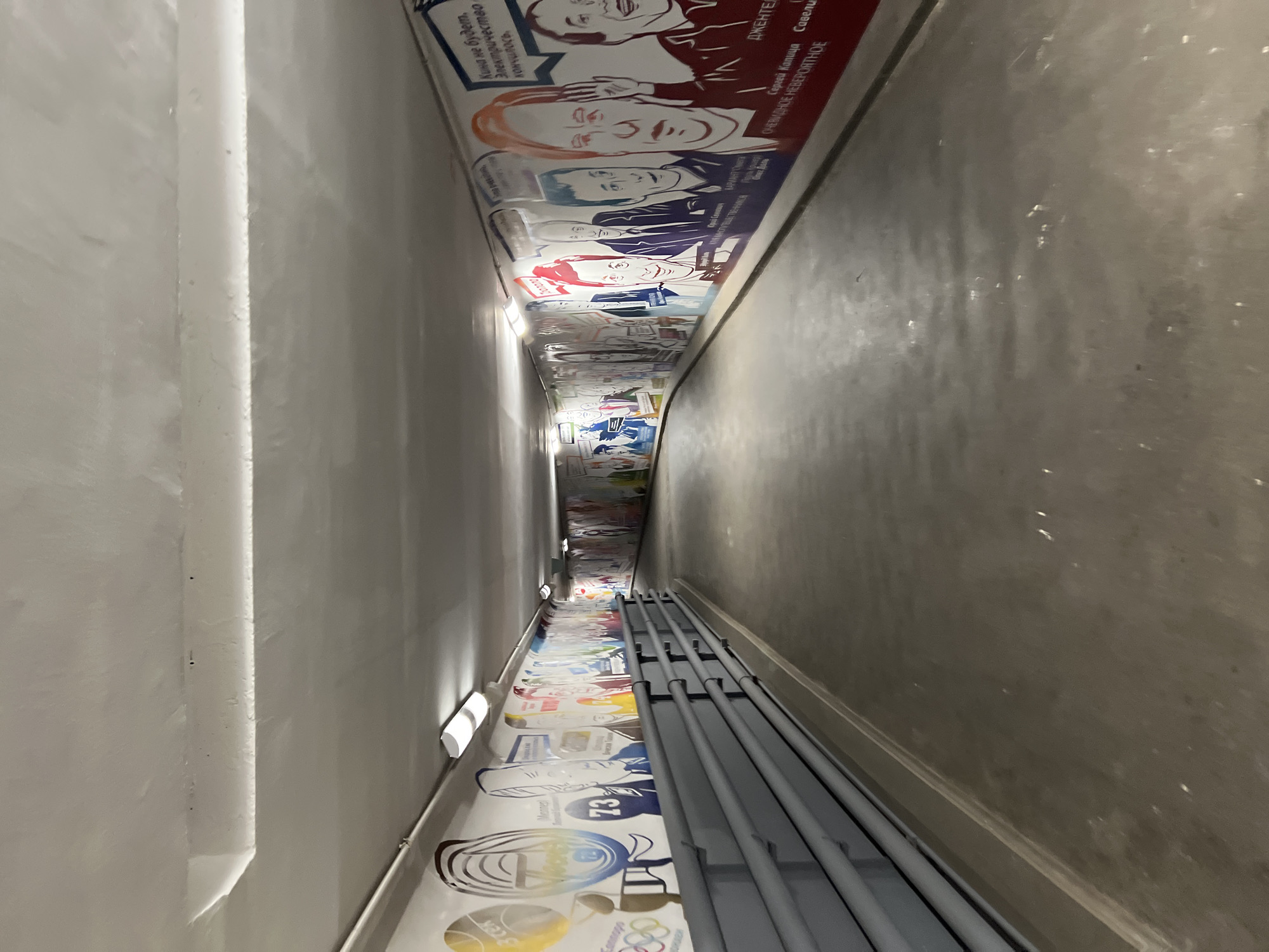 The image depicts a long, narrow, slightly curving corridor or underpass, viewed from a high angle looking down its length. The ceiling is white, and the left wall is adorned with a continuous mural featuring colorful, stylized, and often caricatured portraits of various individuals, primarily men. These murals appear to celebrate famous figures and moments from Soviet/Russian culture and sports.
Visible text on the murals includes the famous Russian cinematic quote: "Кина не будет, Электричество кончилось." (There will be no cinema, electricity has run out.), prominently displayed next to a portrait. Other text mentions "ДЖЕНТЕЛЬМЕНЫ" (Gentlemen) and names such as "Горий Капуша Савелий" (likely referring to characters/actors from the film "Gentlemen of Fortune"), "АРКАДИЙ РАЙКИН," and "АЛЕКСАНДР ШИРВИНДТ," both renowned Soviet/Russian actors and comedians. Further down the mural, elements like "73" and the Olympic rings symbol, possibly accompanied by "Монреаль" (Montreal), suggest a connection to sports history, possibly the 1976 Montreal Olympics.
The right wall of the corridor is plain, made of a darker grey concrete or painted surface. Along the lower section of this wall, several grey pipes run horizontally. The corridor is lit by linear, modern light fixtures mounted on the left wall, casting illumination over the murals. The floor is also a plain grey surface. The overall impression is that of a public pedestrian passage in Khimki, Russia, designed as a commemorative space celebrating cultural and sporting achievements. No direct activity is taking place within the scene.