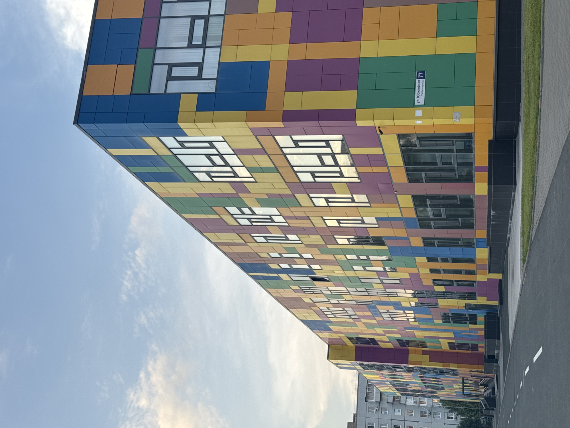 The image shows a brightly colored, modern building with a facade composed of a grid of various colored squares and rectangles in shades of blue, yellow, orange, green, and purple. The building has numerous windows, some of which are large and reflective, showing the sky. A street sign is visible on the building's exterior, reading "ул. Юбилейная 77" and "Yubileynaya St. 77". In the background, to the left, is a more traditional grey apartment building. The scene is an exterior shot, likely taken during the day with a cloudy sky. There are no people visible in the image. The ground in the foreground is paved, with a white line marking a road.