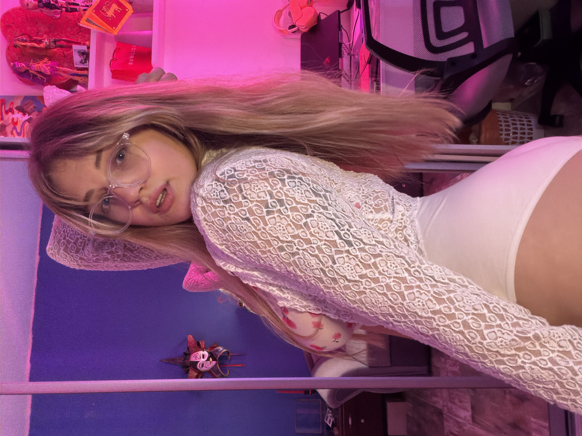 The image depicts a young woman with blonde hair wearing glasses, a white lace top, and a white bodysuit. She is lying on her side, resting her head on her hand. Behind her is a purple wall with a decorative mask. The background includes a shelf with various items, including books and other decorative objects, and a computer chair. The lighting in the room is a mix of pink and purple. There is no evidence to support this photo being taken in the city of Большая Шильна, Russia.