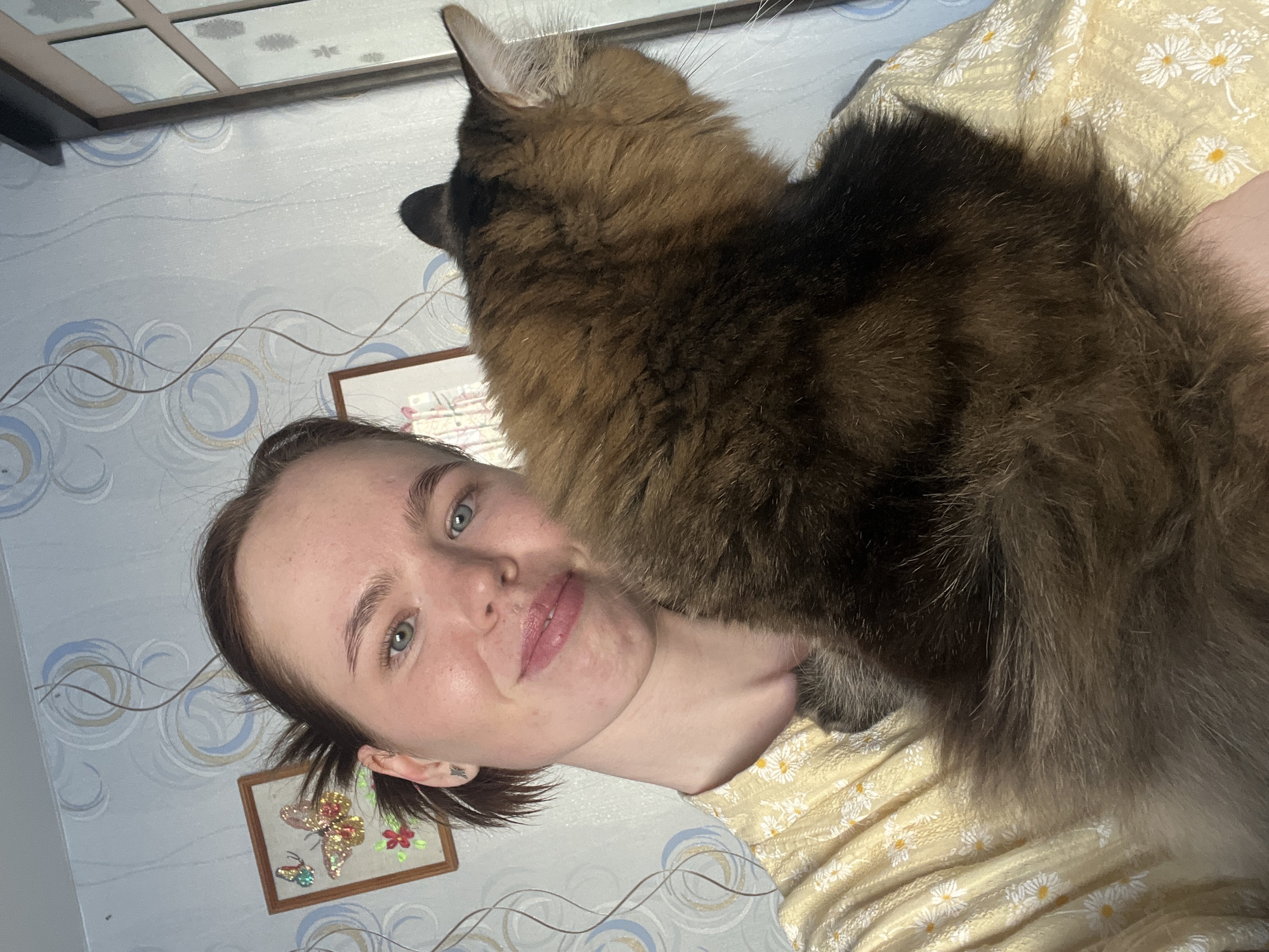 A woman is lying down with a large brown and black cat on top of her. The woman has fair skin, short brown hair, and green eyes. She is wearing a light pink lip color and appears to be smiling slightly. The cat is large and fluffy, with brown and black fur. The woman is lying on a yellow and white floral-patterned bedspread. The background consists of a wall with a wallpaper pattern of blue and white circles and a framed picture of a butterfly and flowers. The location is likely inside a home in the city of Chaikovsky, Russia, but this cannot be confirmed from the image alone.