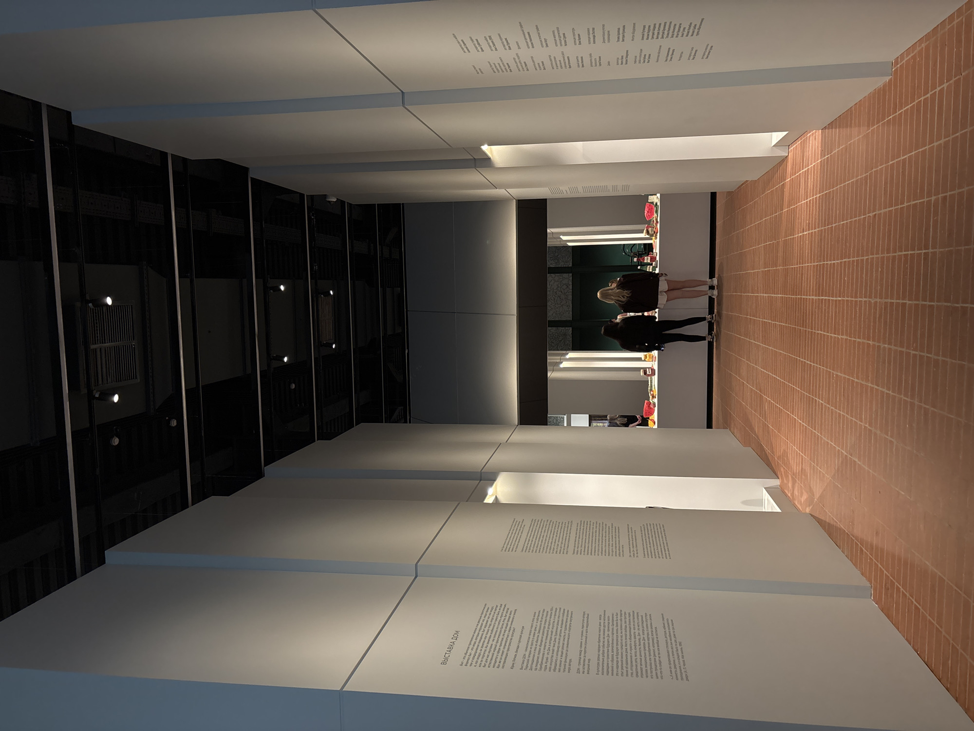 Here's a concise description of the image:
The image is an interior shot of a museum or gallery space, likely in Perm, Russia, judging by the text displayed. The space features a corridor with white walls, some of which are inscribed with text in what appears to be Russian. The floor is tiled in a red-brick pattern. Two people stand further down the corridor, looking at items on display. The lighting is subdued, casting shadows and highlighting the architectural elements. One side of the corridor is made of mirrored panels.