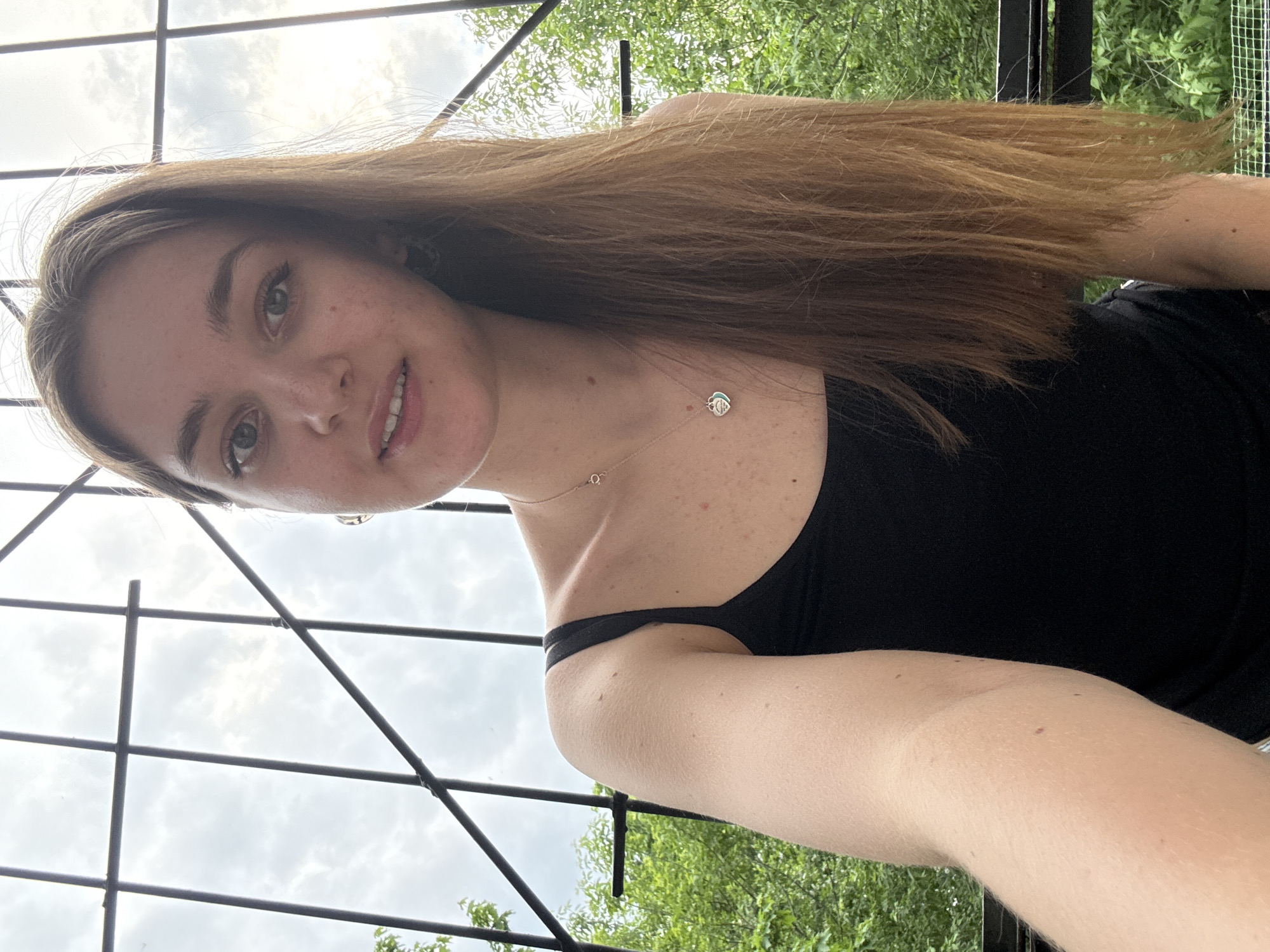 A young woman is taking a selfie. She is wearing a black tank top and a necklace. She has long brown hair. The background consists of a metal structure, likely part of an outdoor setting, with trees and sky visible through the structure. The exact location in Perm, Russia is not determinable from the image.