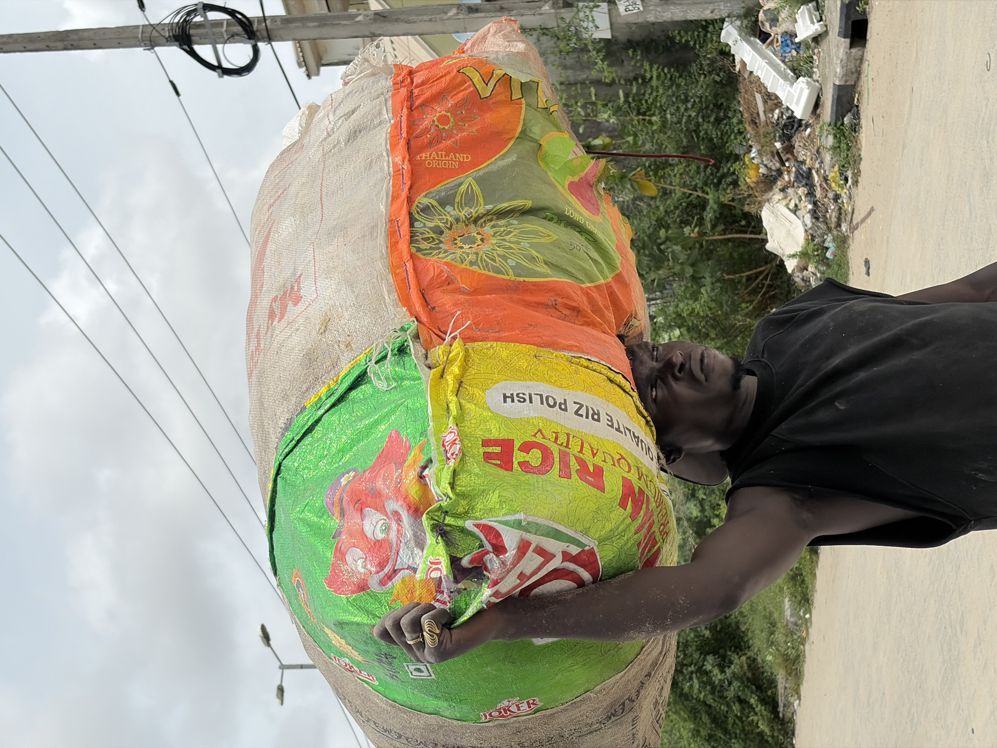 An outdoor, eye-level shot captures a man carrying an exceptionally large and heavy load of sacks on his head, likely in Igboefon, Nigeria. The man, with dark skin, wears a simple black t-shirt and has a focused, slightly upward gaze, indicating the effort involved in balancing the substantial weight. A gold-toned ring is visible on his right hand, which is lightly gripping one of the sacks.
The load consists of several layered bags. The most prominent is a bright green sack printed with a large, vibrant red cartoon character, possibly a pig or boar, and white text that includes "PREMIUM QUALITY RIZ POLISH" (partially inverted) and the brand "JOKER." Below this, an orange and green sack with a floral design is visible, featuring text like "THAILAND ORIGIN" and "LONG GRAN" (likely "LONG GRAIN"). The outermost layer appears to be a coarse, light-colored woven material, possibly hessian or polypropylene.
The setting suggests a semi-urban or rural environment. In the background, power lines stretch across a cloudy, bright sky. A utility pole with a cluster of black cables is visible. To the right, lush green foliage and scattered debris, including white polystyrene pieces and other waste, line the edge of a dirt or sandy road with a concrete curb. The overall scene depicts a common sight of manual labor and goods transportation in many parts of the world, highlighting resilience and daily life in the region. The time of day appears to be daytime, possibly overcast or bright, but not direct harsh sunlight.