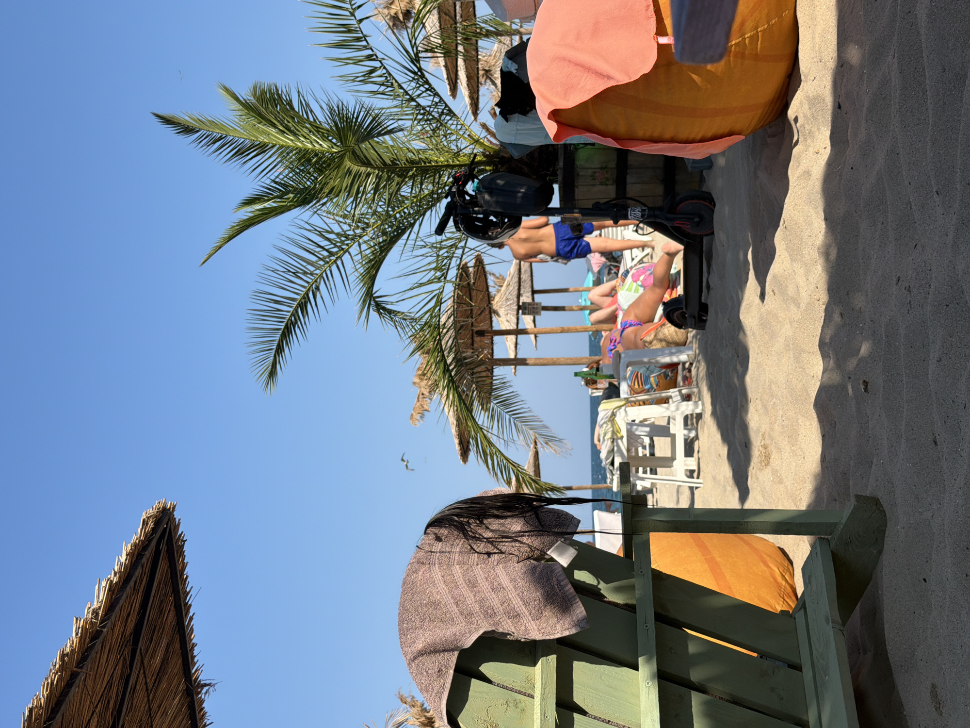 The image depicts a sunny day at a beach, likely in Varna, Bulgaria, given the context provided. In the foreground, a green Adirondack chair is positioned on sandy ground, with a brown towel draped over its back, partially obscuring long, dark hair hanging from it. Adjacent to the chair, a large, round, orange beanbag-like object covered with a pink towel is visible.
Further into the scene, beach umbrellas with straw canopies provide shade over white lounge chairs. A palm tree stands to the right, its fronds reaching towards the clear blue sky. Near the palm tree, a black electric scooter is parked, with a black helmet attached to its handlebars. A wooden barrel, possibly part of a stand or decoration, is located behind the scooter.
In the background, several people are visible relaxing on lounge chairs. One person, wearing blue swim trunks, stands near the palm tree. Another person, lying on a lounge chair, is partially visible. The sea and horizon are discernible in the distance, suggesting a coastal location. The overall atmosphere is one of leisure and a typical summer beach day.