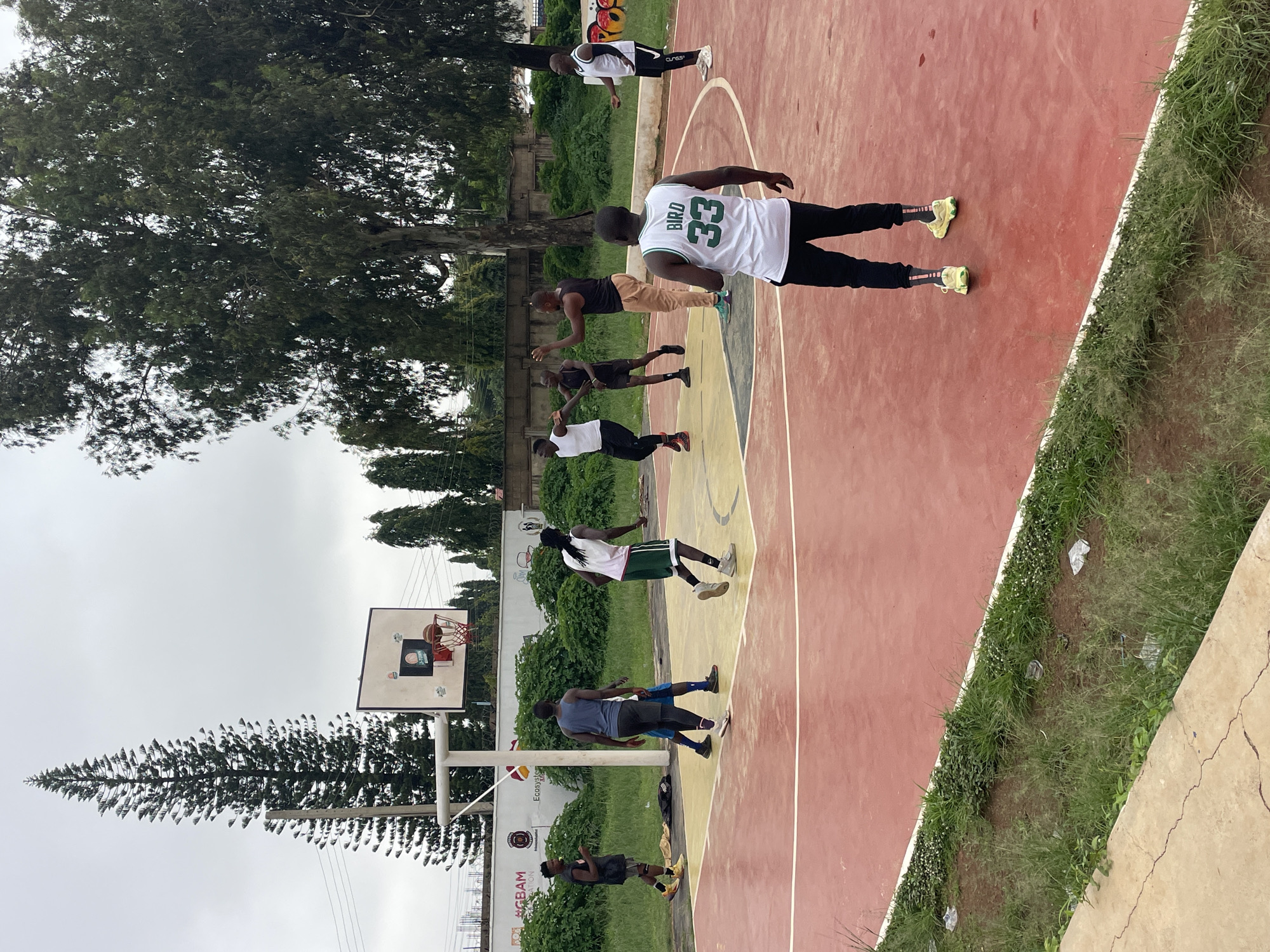 A group of young African men are playing basketball on an outdoor court under a cloudy sky in Gura Top, Nigeria. The court is red and yellow with white lines. A basketball hoop is visible in the background. Several trees surround the court, and a tall pine tree is on the left side of the image. Some of the men are wearing athletic clothing, and one is wearing a white jersey with the name "BIRD" and the number "33" on the back. The men are actively engaged in the game, with some dribbling the ball and others positioned to receive a pass. The environment appears to be somewhat unkempt, with grass and scattered debris around the court.