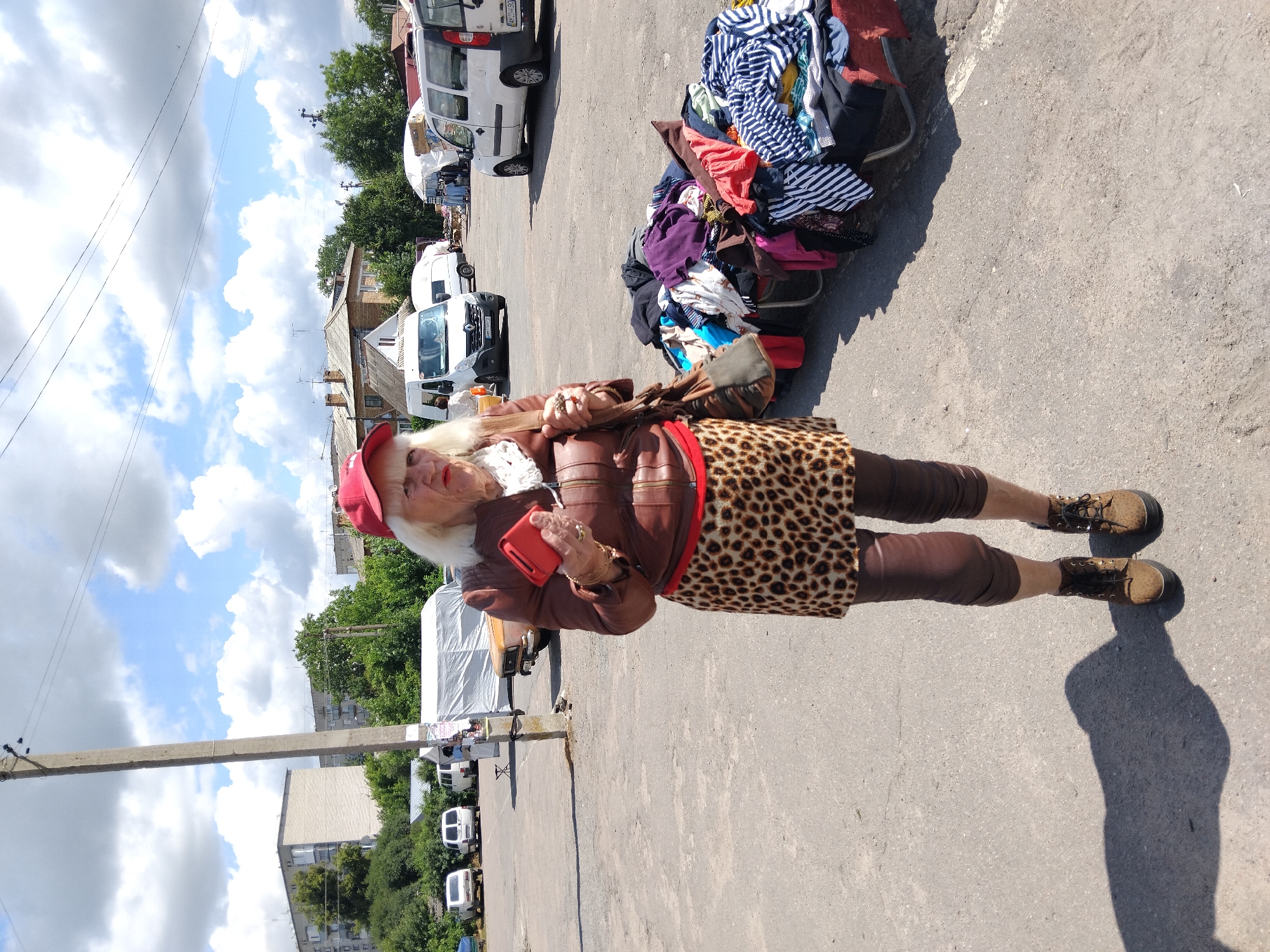An elderly Caucasian woman wearing a red baseball cap, a brown leather jacket, a leopard print skirt, and brown leggings stands in a paved outdoor market area. She holds a red smartphone in her right hand and a dark brown purse on her left shoulder. In the background, several white vans and a tan car are parked. To the right of the woman, a large pile of folded and draped clothing is displayed on a cart. The environment appears to be a sunny day with scattered clouds in the sky. This scene is located in Chudniv, Ukraine.