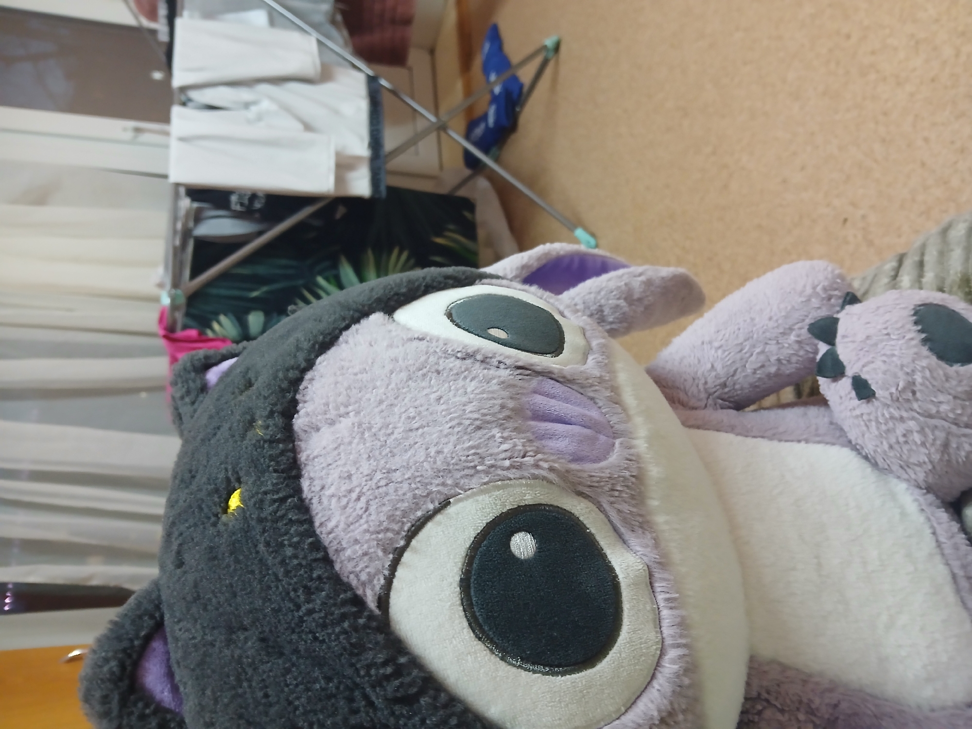 The image shows a close-up of a plush toy, likely a stuffed animal. The toy is predominantly purple and white, with a large, black eye and purple ears. There appears to be a black hooded section on the head of the toy.
The background includes a clothes drying rack with white clothes, a blue chair, and a beige wall. The scene appears to be indoors.
There is no information to place the location in the city of Chaikovsky, Russia.
