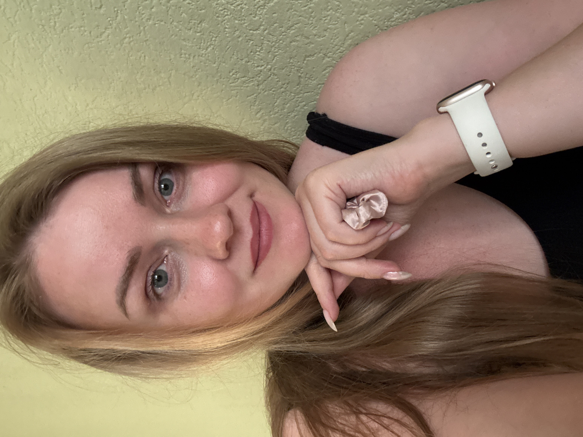 A young woman with blonde hair is captured in a close-up selfie. She is leaning her head on her hand, with her chin resting on her knuckles. Her eyes are open and looking towards the camera. Her lips are slightly parted, and she appears to be smiling subtly. She is wearing a black top and a white watch on her left wrist. Her fingernails are long and painted. The background is a textured, pale green wall. The image was taken indoors. The location is not specified in Chaikovsky, Russia.