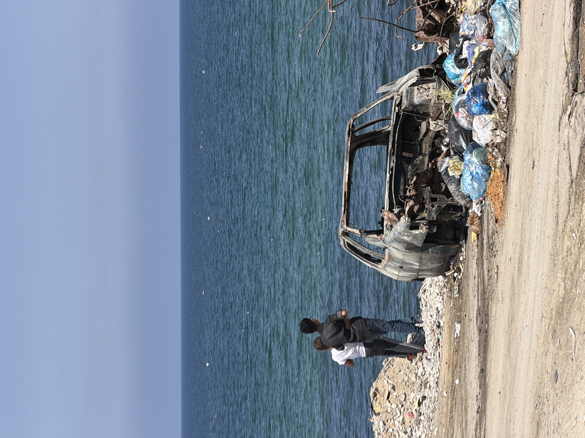 Two young males, one with his arm around the other, stand near the shore, gazing out at the ocean. They are positioned to the left of a burnt-out vehicle, which sits partially submerged in a pile of trash. The scene is located in an urban environment by the sea, evidenced by the presence of a shoreline, garbage, and a destroyed car. The sky is clear and blue, and the ocean is a deep blue.