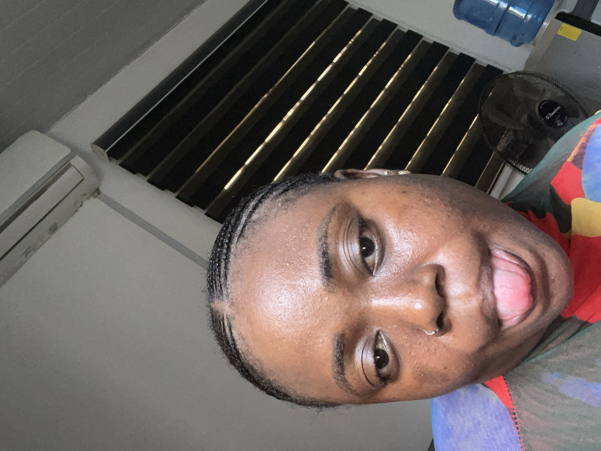 A close-up, tilted view captures a dark-skinned woman with braided hair, a nose ring, and an open mouth. Her face is the primary focus of the image. The background includes horizontal blinds with light filtering through, an air conditioning unit, a fan, and a water dispenser. The location is inferred to be in Abuja, Nigeria, based on the prompt. The woman is wearing a multicolored garment, and the scene appears to be indoors.