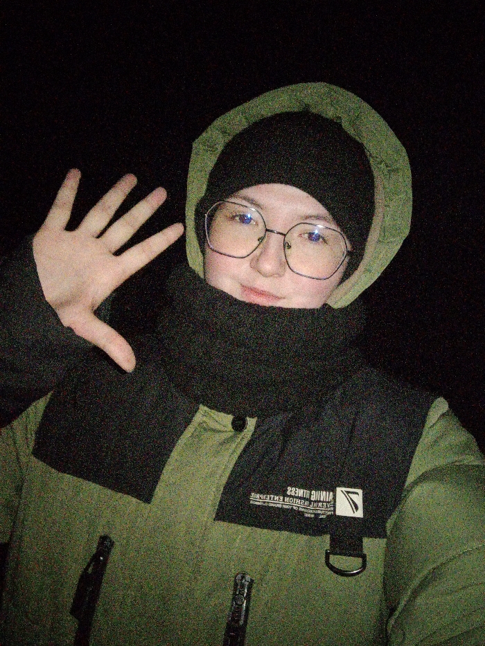 A nighttime, portrait-oriented image shows a single individual from the chest up, against a completely dark, featureless background, located in Старый Шагирт, Russia. The subject wears hexagonal eyeglasses, a black beanie, an olive-green hooded jacket with the hood up, and a black neck gaiter covering the lower face and neck. The individual's right hand is raised to the left of their head, palm forward with fingers splayed, in a gesture. A slight smile is present. The jacket features black shoulder and upper chest panels. Reversed white text reading "VIKING FITNESS" and a logo are visible on the right chest panel, along with a black D-ring attachment. The image exhibits significant digital noise, consistent with low-light conditions and flash illumination.