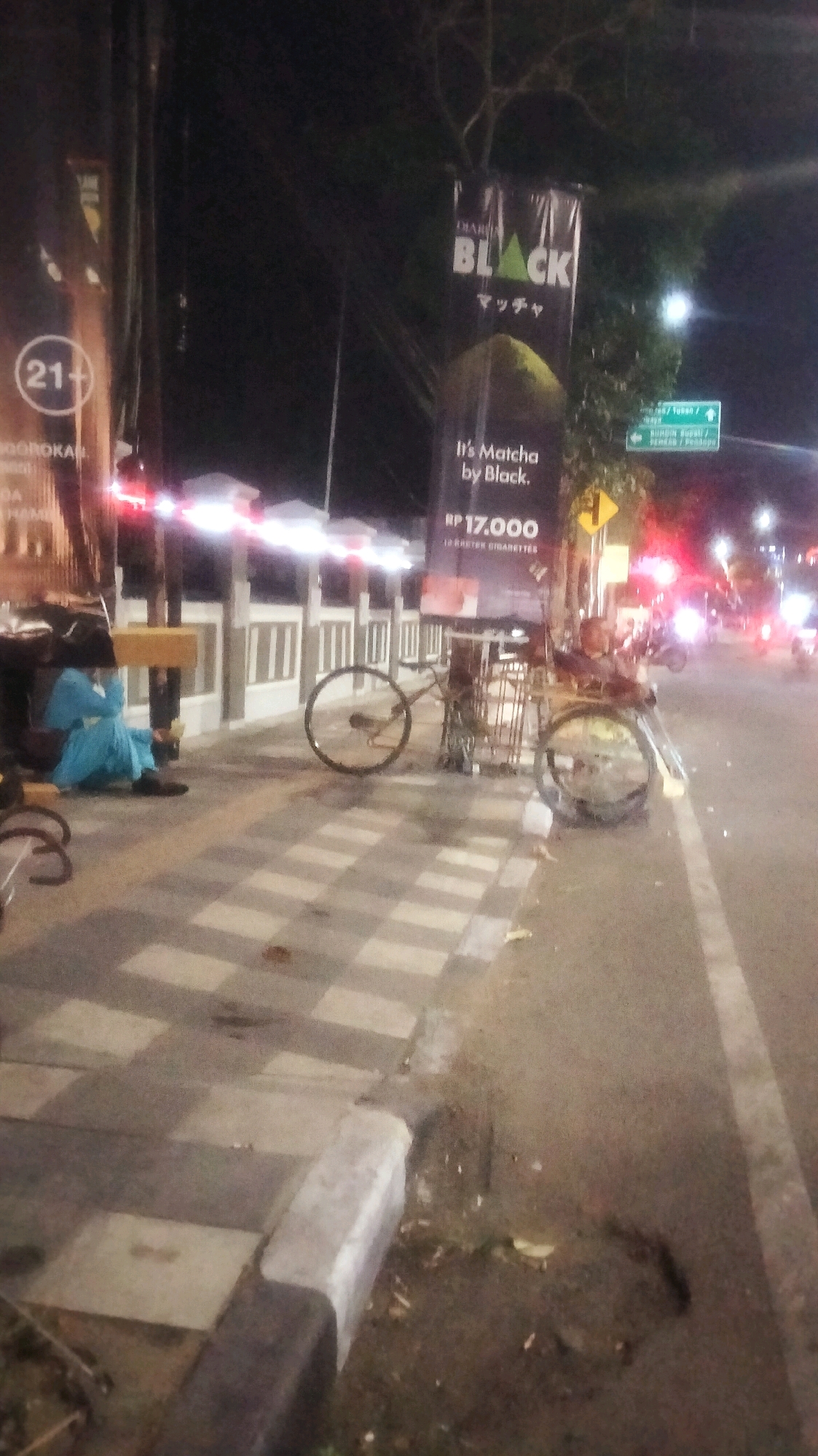 The image shows a street scene at night in Bojonegoro, Indonesia. On the left, a person wearing a blue outfit sits on the sidewalk next to a wooden bench. Behind them, a banner displays "DIARI BLACK Matcha" and a price of "RP 17.000". To the right of the person, a bicycle is parked on the sidewalk. The sidewalk is paved with a checkered pattern. Further down the street, there are lights and traffic, suggesting a busy urban area. A green street sign indicates directions to "Tuban" and "Cepu".