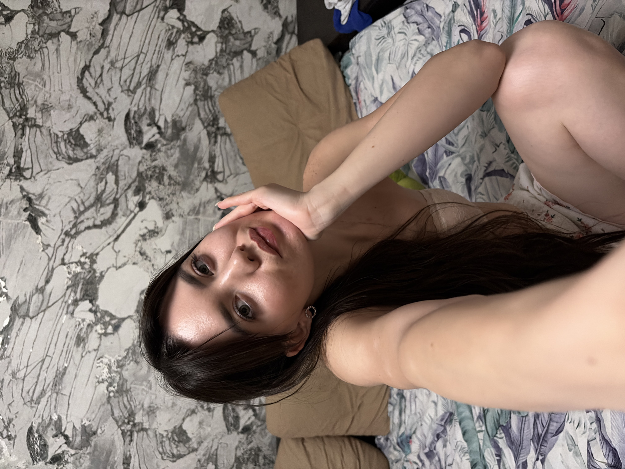 A young woman lies on a bed in a room. The wall has a marble-like wallpaper. She is holding her hand near her face. She has long dark hair and wears a small earring. The bedding has a floral and feather print. The location is likely Chaikovsky, Russia, but this cannot be confirmed without additional evidence.