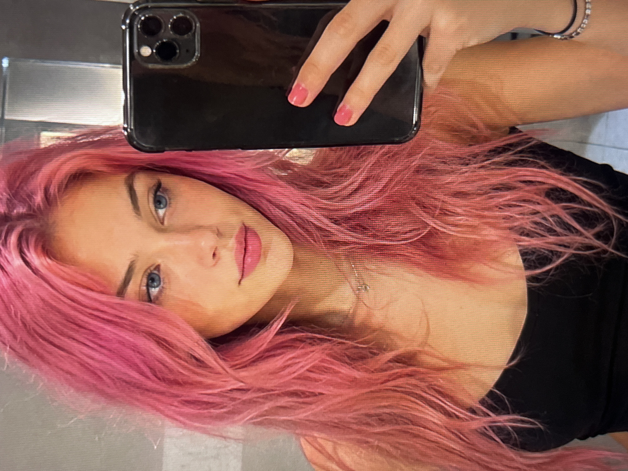 The image is a close-up, head-and-shoulders selfie of a young woman with bright pink hair. She is holding a black smartphone in her right hand, angled towards herself, indicating she is taking the picture. Her left hand is visible, resting on the side of the phone. The woman has blue eyes and is wearing a black tank top. A delicate silver necklace is visible around her neck. Her fingernails are painted a bright pink. The background appears to be a room with a neutral-colored wall, possibly a bathroom due to the lighting and the reflective surface visible behind her. The image has a slightly grainy texture, suggesting it was taken with a smartphone camera. There are no clear indicators of a specific city or location within a city.