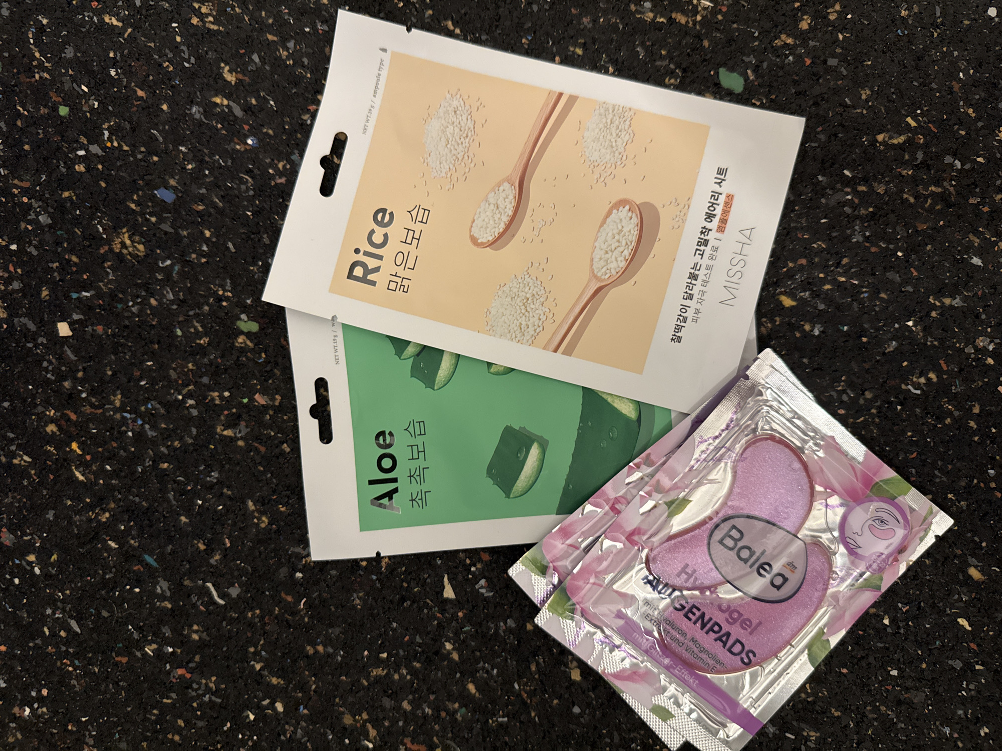 An overhead shot shows three beauty products placed on a dark, speckled surface. The products are arranged in a loose triangle. The bottom left object is a silver foil packet containing two pink, crescent-shaped under-eye masks. The packet is adorned with floral imagery and has text in English and German. To the right of the packet is a light green package with the word "Aloe" in large, black font, followed by Korean text and an illustration of aloe vera slices. Further to the right is a light peach-colored package labeled "Rice" in large, black font, also with Korean text and images of spoons filled with rice. The background surface appears to be a textured, composite material with flecks of various colors, resembling recycled rubber flooring.