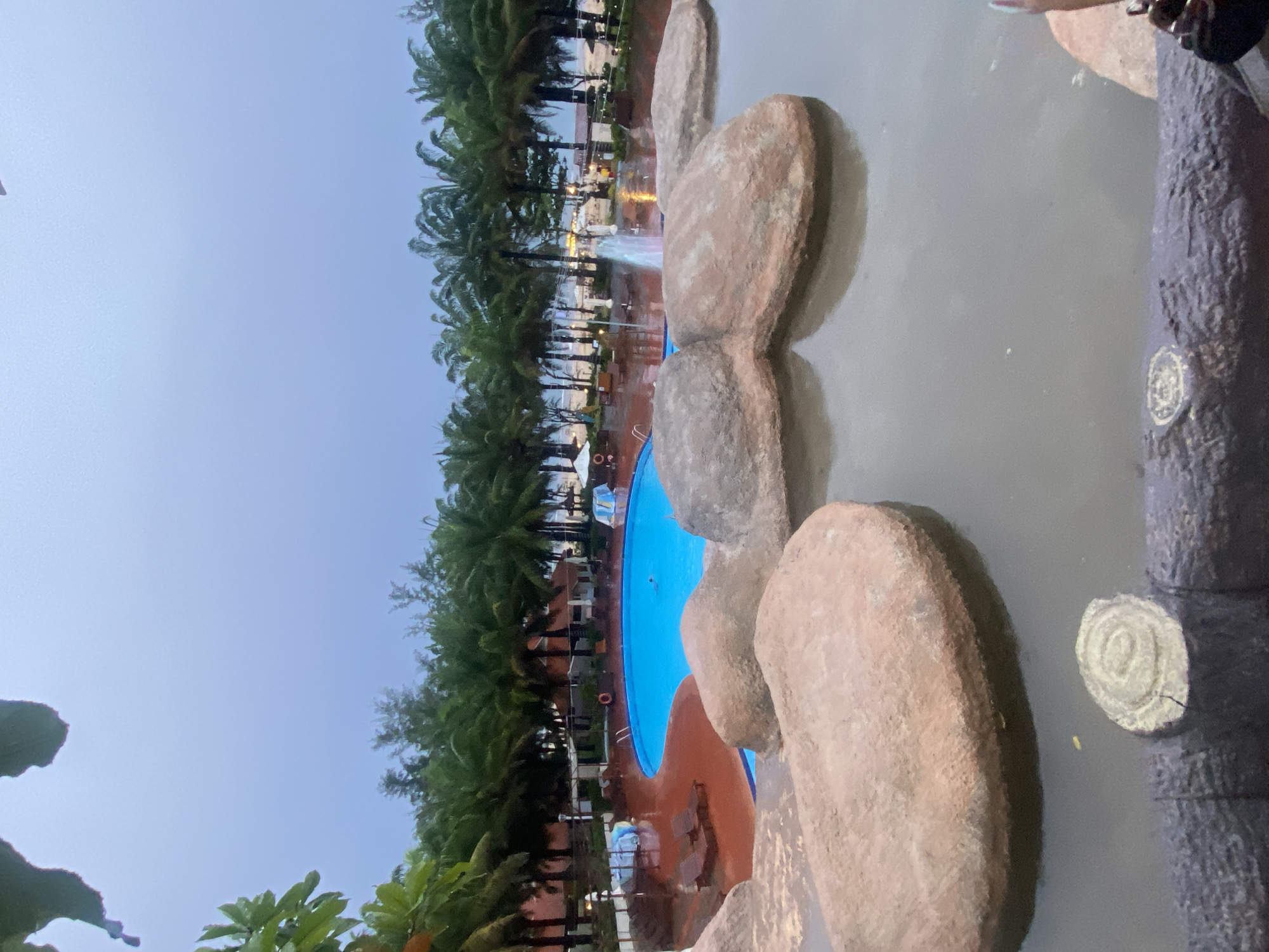 This is an outdoor scene in Phu Thuy Ward, Vietnam, likely at a resort or hotel. The photograph is taken from an elevated position, possibly from within a building or structure.
The main subject is a swimming pool, partially visible on the left, with a blue interior and a brown surround. Palm trees line the perimeter of the pool area, stretching into the background. In the distance, buildings and lights suggest a developed area, potentially the resort itself or a nearby town.
Dominating the foreground on the right side are large, artificial rock formations, appearing to be part of the landscaping or pool design. The foreground is slightly out of focus.
The sky is overcast, indicating it's either early morning or late evening. The overall atmosphere seems tranquil.