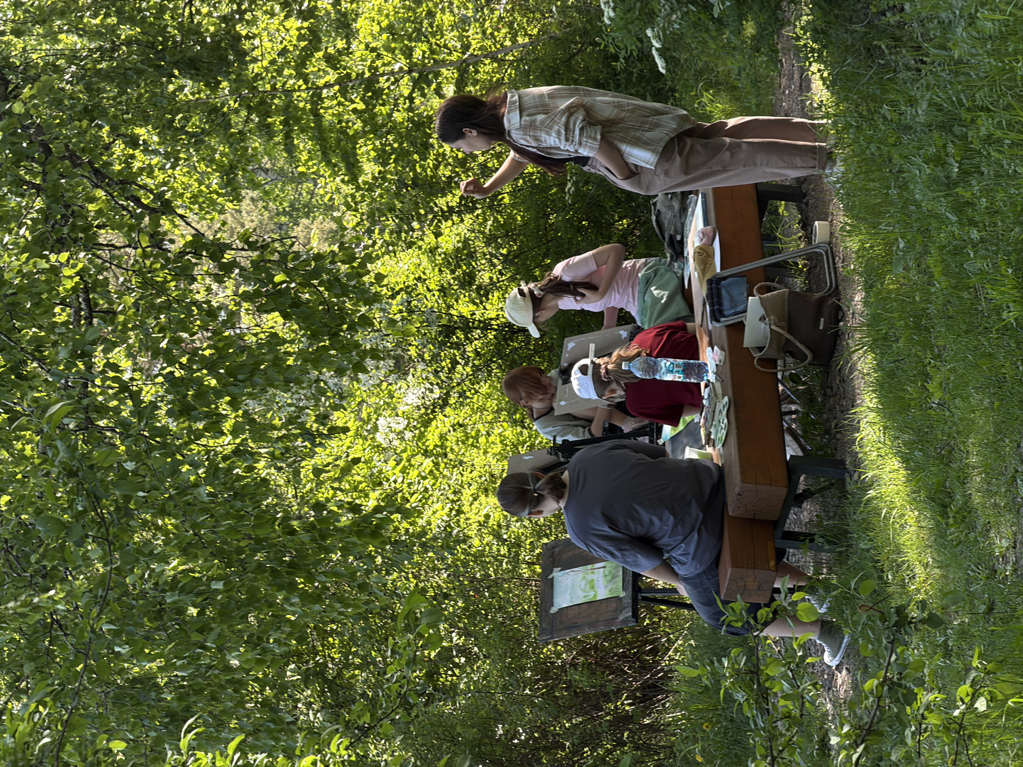 The image depicts an outdoor gathering, likely an art or nature study class, set in a lush green natural environment, possibly a park or wooded area. Five individuals are visible, engaged in an activity around a long wooden bench or table structure.
On the left side of the corrected image, one person stands, wearing a plaid shirt and light pants. They are leaning forward, with an arm extended, appearing to instruct, observe, or demonstrate something to the seated participants.
Four other individuals are seated along the wooden structure. From left to right:
1. A person in a pink shirt and white baseball cap, looking down at their work.
2. A person in a red shirt. A clear plastic water bottle with a blue cap and a label is placed on the table in front of them.
3. Another person, partially obscured by the individual in the red shirt, wearing a lighter-colored top.
4. A person in a dark grey short-sleeved top, facing away from the viewer. They are focused on an easel or drawing board placed on the ground in front of them, which holds what appears to be a light-colored sketch or painting.
Various items associated with outdoor activities and art are scattered around the bench, including bags (one woven, one backpack-like), a tablet or small screen, and other supplies.
The scene is surrounded by abundant, vibrant green foliage from trees and bushes, indicating a natural setting. Bright natural sunlight filters through the leaves, creating dappled light and shadows on the ground and participants. The weather appears clear and sunny, suggesting a warm day. No discernible text is visible on any objects, apart from the unreadable label on the water bottle. The overall atmosphere is one of focused activity in a peaceful outdoor environment. The location is contextualized as Krasnoyarsk, Russia.
