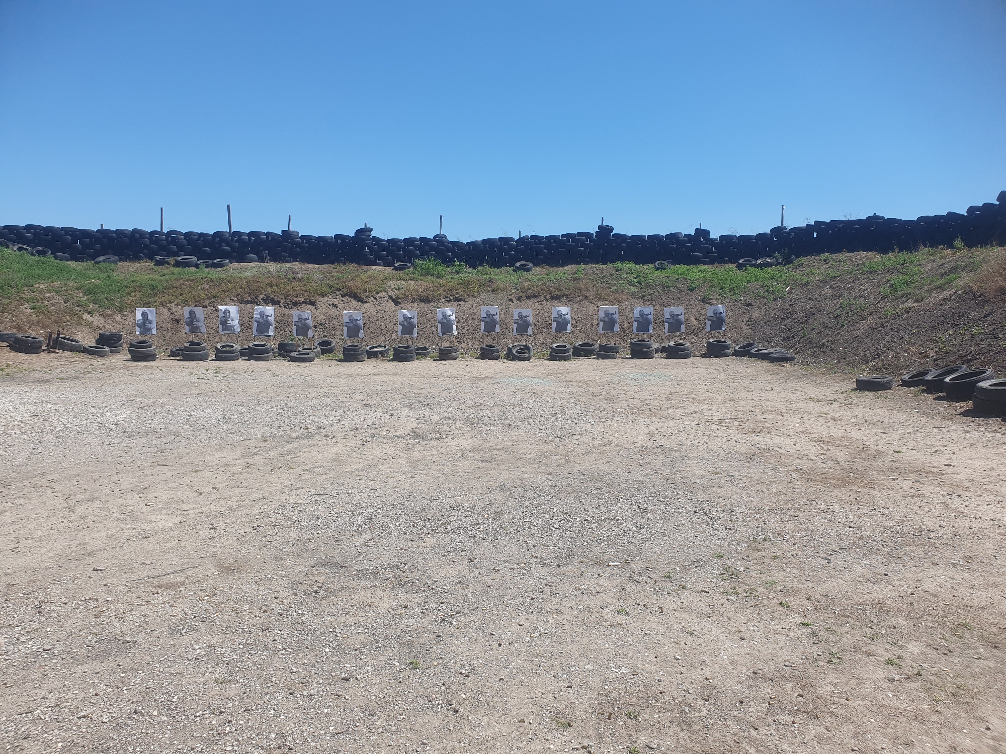 The image shows an outdoor shooting range located in Войводино, Bulgaria. The range consists of a gravel area with a line of targets arranged in a row. The targets are humanoid silhouettes mounted on what appears to be tires. A barrier constructed of stacked tires and earth is present behind the targets, serving as a backstop. The sky is clear and blue.