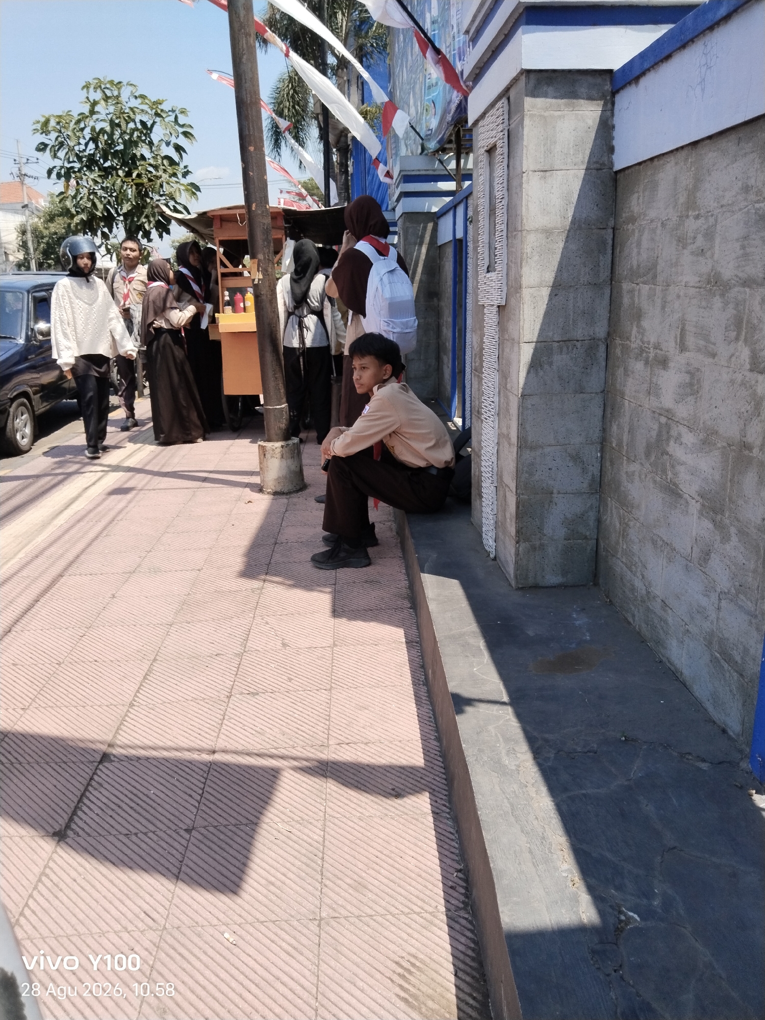 The image depicts a sunny outdoor scene in Situbondo, Indonesia, likely in front of a school or public building. A group of students in school uniforms are gathered near a food stall. One student, a young boy wearing a tan shirt and dark pants, is sitting on a ledge and looking away from the camera. Another student, a girl in a white sweater and black pants with a helmet, is standing near a black car. Several other students in similar uniforms are visible, some interacting near the food stall. Red and white Indonesian flags are strung across poles in the background. The ground is paved with reddish-pink tiles and a dark concrete walkway. The wall of a building, constructed from grey concrete blocks, forms the backdrop on the right side of the image. A timestamp in the lower left corner indicates "vivo Y100 28 Agu 2026, 10.58".