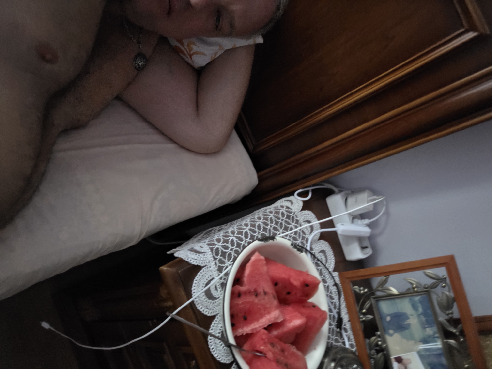 A first-person perspective shot captures a man lying on his side in bed, with his left arm propped behind his head. He is wearing a necklace. On a bedside table next to the bed, a bowl of sliced watermelon sits atop a doily. A power strip with two plugs and a phone charger is also visible on the table. In the background, a framed picture hangs on the wall, displaying two individuals. The scene appears to be indoors, likely in a residential setting in Batumi, Georgia.