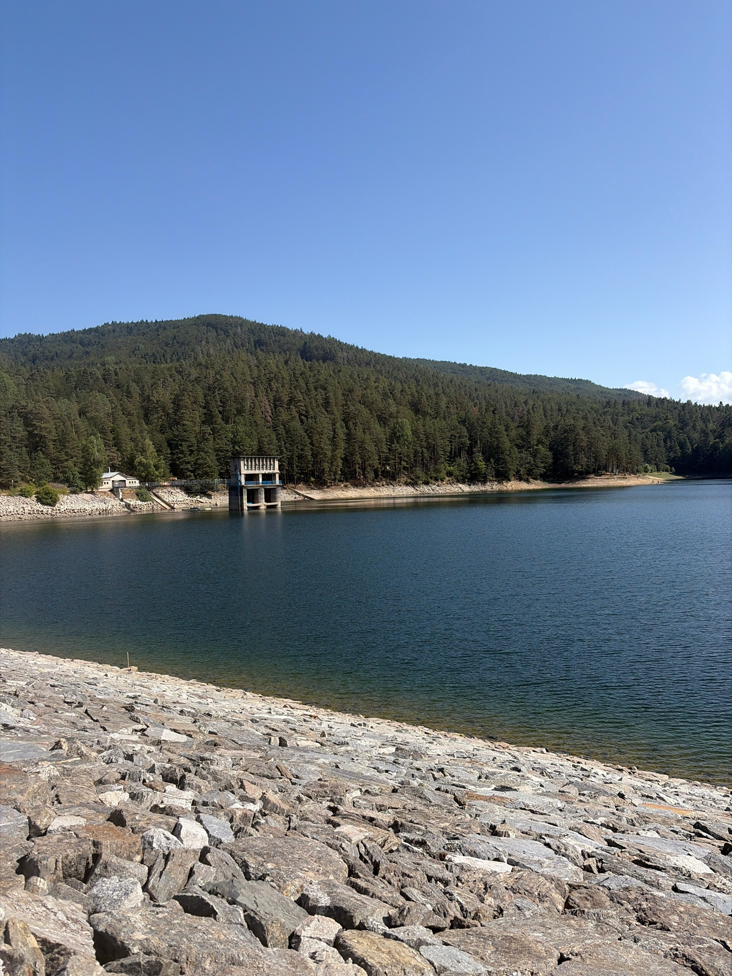 The image depicts a wide shot of a reservoir, likely the Sv. Konstantin Reservoir, near the city of Sv. Konstantin, Bulgaria. The foreground is dominated by a slope covered in large, grey, irregular stones, suggesting a constructed dam or embankment. The dark blue water of the reservoir stretches across the mid-ground, reflecting the clear, bright blue sky above.
Along the far bank of the reservoir, a dense, green pine forest covers a gently sloping hill that rises to form a mountain range in the background. The trees appear healthy and vibrant, indicating a natural, wooded environment.
On the left side of the reservoir, near the edge of the water and at the base of the forest, there are a few man-made structures. Closest to the water's edge is a small, light-colored building with a prominent roof. Further inland and slightly to the right of this building, a more substantial concrete structure with multiple openings and a blue-railed balcony stands adjacent to the water. This structure appears to be part of the reservoir's infrastructure, possibly a water intake tower or control building. A walkway or bridge connects this structure to the shore.
The overall scene conveys a sense of natural beauty and functional engineering, with the reservoir serving as a focal point within a serene, forested landscape. The lighting suggests it is daytime, likely mid-morning or early afternoon, given the clear sky and the way light falls on the scene. No people are clearly visible in the image.
