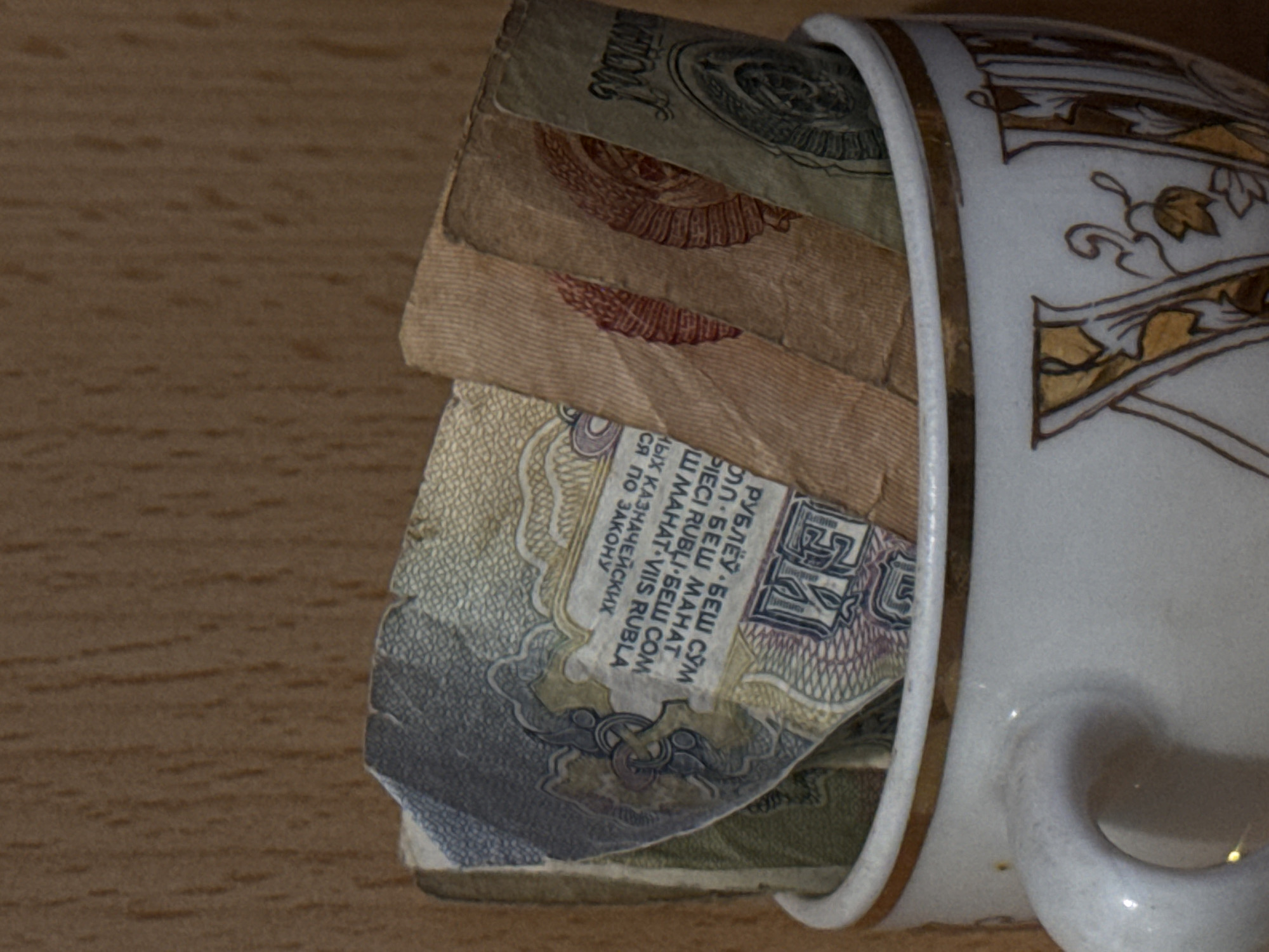 A porcelain cup with a gold leaf pattern on the side is filled with several Soviet-era banknotes. The top note is faded and worn, with a visible denomination of "100". The note underneath has a prominent emblem of a hammer and sickle. The note below that has the text "РУБЛЁЎ БЕШ СЎМ ...". The cup is placed on a wooden surface. There are no people visible in the image. The location is not inferable from the image.
