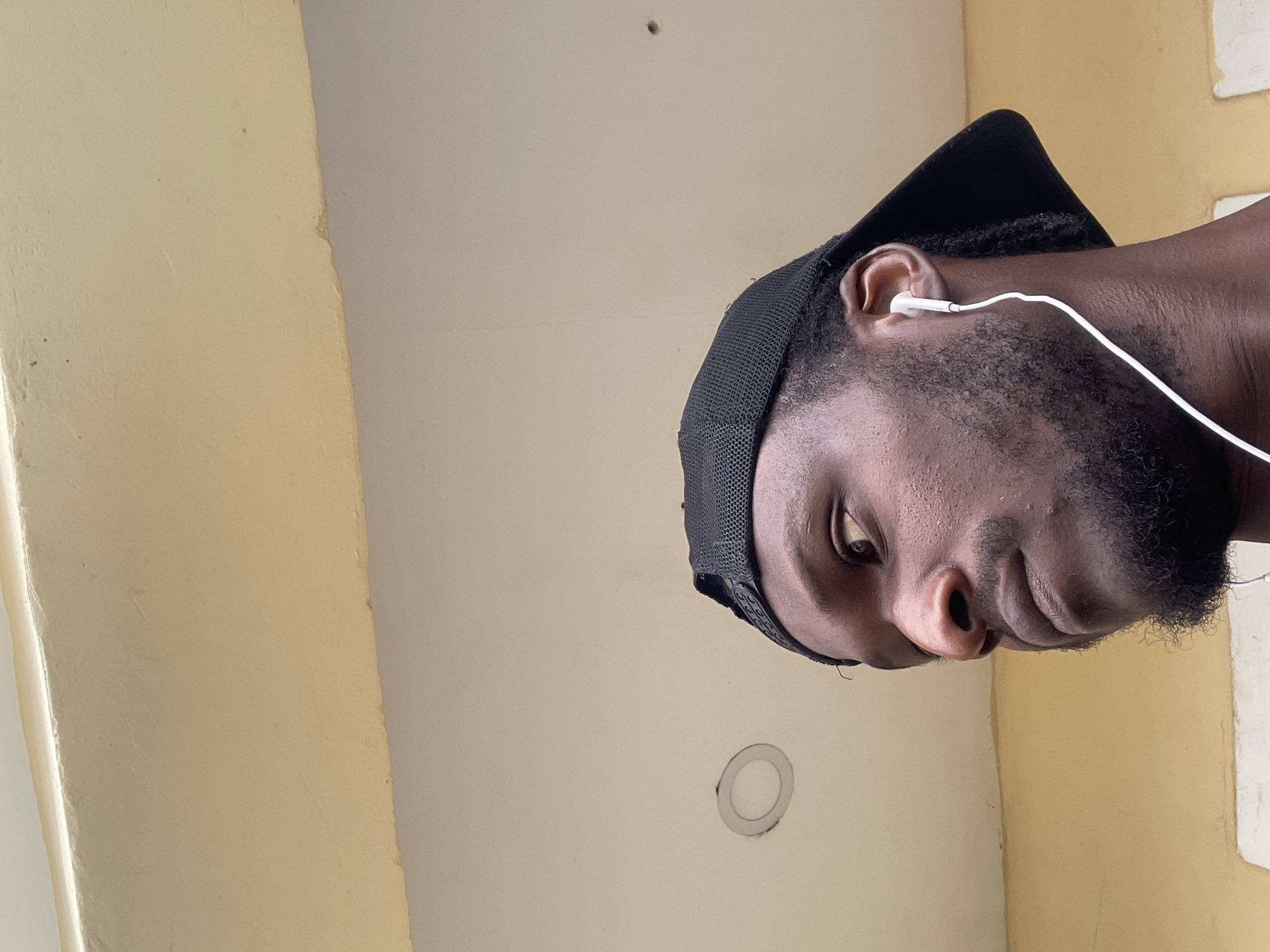 A close-up, low-angle shot shows the left profile of a Black man wearing a black baseball cap and white earbuds. He has dark skin, a short beard, and is looking to his left. The background consists of a beige ceiling with a circular light fixture and a yellow wall. The image was taken outdoors in Abuja, Nigeria.