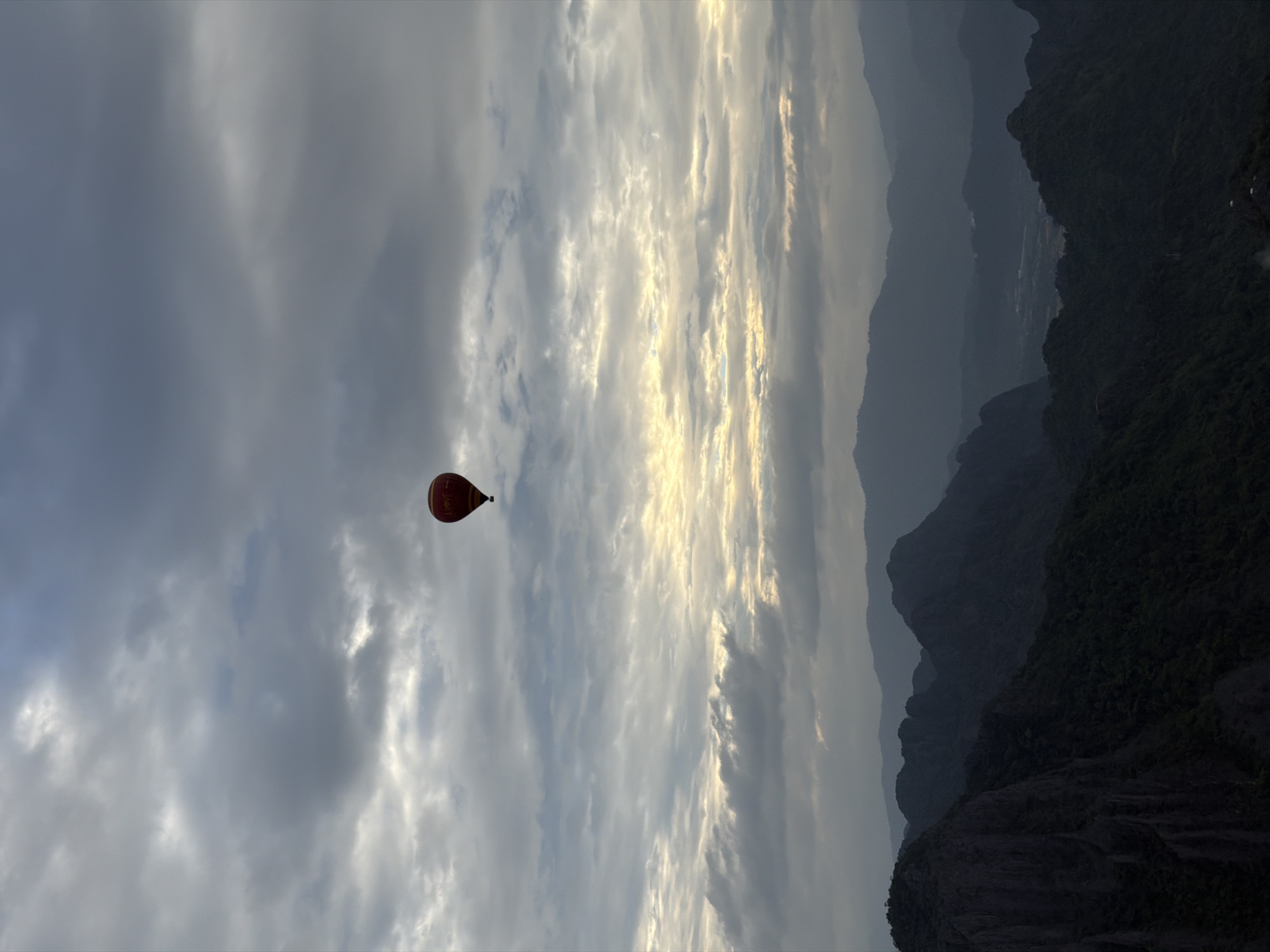 A hot air balloon is suspended in a cloudy sky above a mountain range. The balloon, colored red with yellow lettering, is small in the frame. The mountains below are dark and appear to be covered in dense vegetation. The sky is a mix of gray and white clouds, with some areas of lighter, possibly sunlit, sky. The overall scene suggests an early morning or late afternoon with the sun partially obscured by clouds. The location is Vang Vieng, Laos, known for its karst landscapes and outdoor activities such as hot air ballooning.