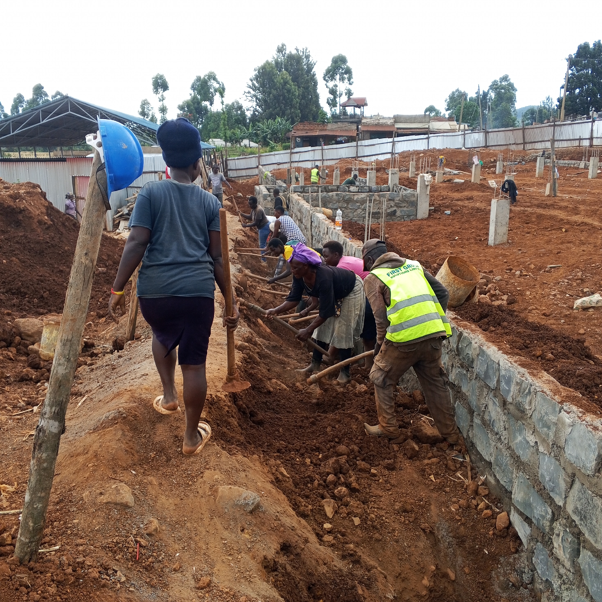 This image captures a vibrant construction site in Borabu Chitago ward, Kenya, teeming with manual labor. The scene is dominated by reddish-brown soil, indicating recent earthwork.
In the foreground, a person walks away from the camera along a dirt path, wearing a blue knit cap and a dark t-shirt, with a blue hard hat hanging on a wooden post beside them. Further along the path, a group of workers is actively engaged in digging a trench. They are using traditional hoes with long wooden handles to break up and move the soil. Most of these workers appear to be women, dressed in everyday clothing.
To the right, partially constructed foundations made of rough-hewn stone blocks are visible, forming the base of a new structure. Numerous concrete pillars rise from the ground, marking the layout of the developing buildings. One worker in a high-visibility yellow vest, bearing the partially visible text "FIRST GROU SOLUTIONS LIMI," is also engaged in the digging.
In the background, more concrete pillars are scattered across the site. Beyond the construction area, a metal fence separates the site from various structures, including buildings with corrugated iron roofs and walls. Lush green trees dot the landscape under what appears to be a bright, possibly overcast, daytime sky. The image emphasizes the intensive manual effort involved in the construction process in this region.