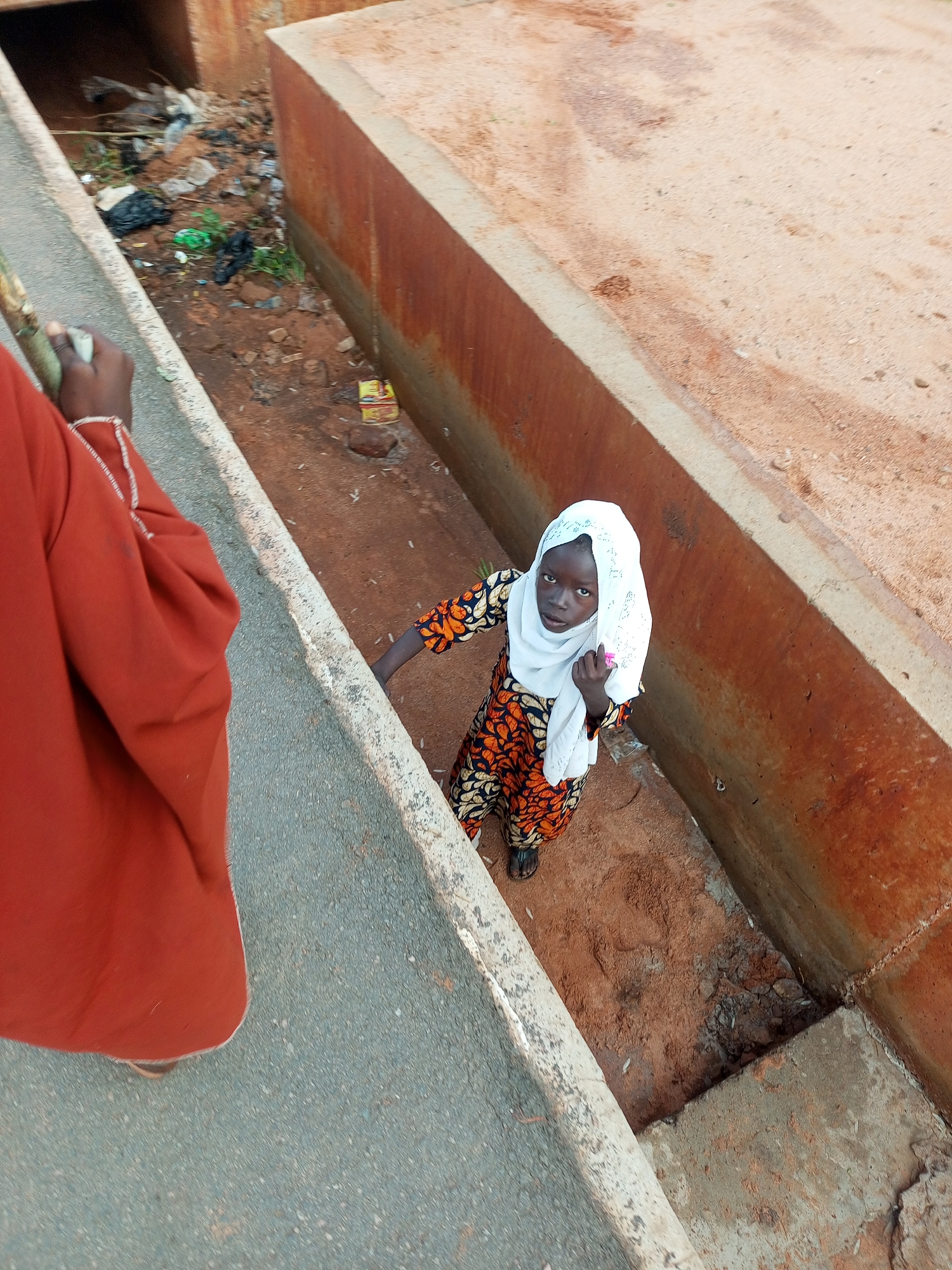 An overhead photograph depicts a scene featuring two individuals, one fully visible and one partially obscured, within an outdoor urban or semi-urban environment in Unknown, Unknown.
In the foreground, the left side of an individual, likely an adult, is visible. This person wears a reddish-orange garment with white stitching details along the shoulder and sleeve. Their left hand is visible, holding a long, light-green fibrous object, possibly a plant stalk or sugarcane, partially wrapped in a light-colored material. This individual is positioned on a dark gray, coarse paved surface, likely a road or pathway, which runs horizontally across the lower left of the frame.
Parallel to the paved surface is an open, unkempt drainage ditch or canal. This ditch features reddish-brown concrete or brick walls and a bottom filled with reddish-brown soil and sand. Standing within this ditch, facing upwards towards the camera, is a young girl. She has dark skin and eyes and a serious expression. She is dressed in a long-sleeved tunic or dress with a prominent orange, black, and white patterned fabric. A white, perforated or patterned headscarf (hijab) covers her head and frames her face. Her feet, clad in dark flip-flops or sandals, are visible at the bottom of the ditch. Her right hand is slightly raised, holding a small, light pink object between her thumb and forefinger.
The bottom of the ditch, particularly in the background, contains scattered refuse including black and green plastic bags, white plastic pieces, crumpled paper, and other debris. Beyond the ditch to the right, a wider expanse of reddish-brown sandy ground is visible. In the far background, the ditch appears to continue under a structure or culvert. The scene is illuminated by daylight, with a top-down perspective.