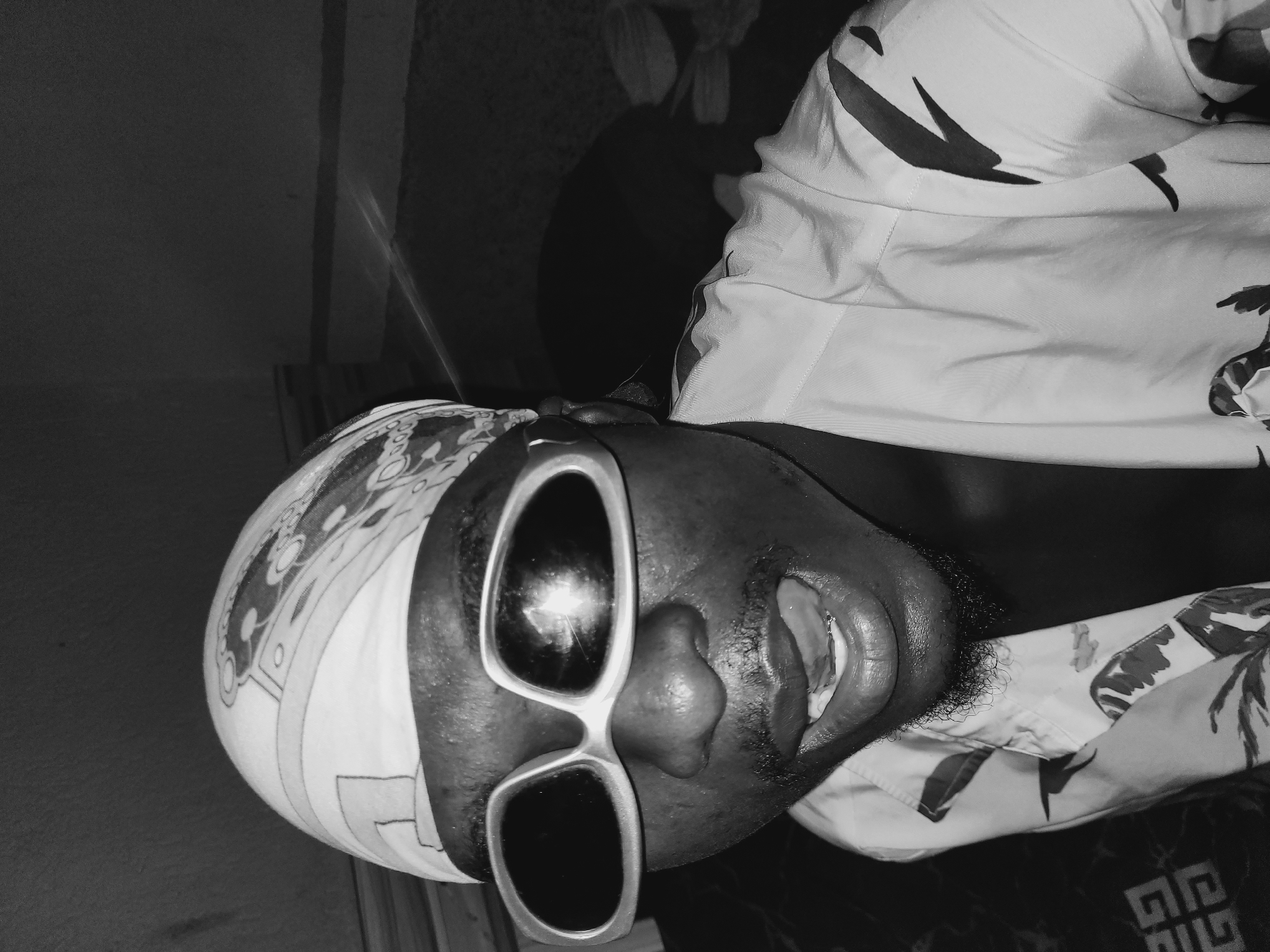 In a close-up, black and white, waist-up shot, a Black male is positioned centrally, looking directly at the camera. He wears a bandana patterned with an intricate design and oversized, futuristic-style sunglasses with thick white rims. His mouth is open, revealing his tongue and teeth, and he has a goatee and mustache. He is dressed in a patterned button-down shirt, possibly with tropical motifs. The background is dark and out of focus, with hints of furniture or a wall behind him. The image appears to be a selfie taken indoors, potentially in Bukuru, Nigeria, given the context.