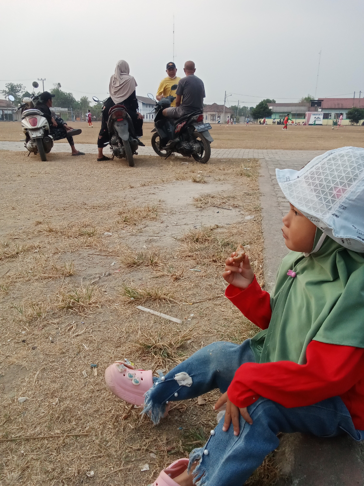 In the city of Toto Katon, Indonesia, this image captures a scene outdoors with a clear sky and a sparse, dry landscape. In the foreground, a young child wearing a light-colored bucket hat, a green outer garment, and a red long-sleeved shirt, sits on a grey rock, holding a small food item in their right hand. The child's blue jeans are ripped at the knee and adorned with pearl-like decorations. They are also wearing pink Crocs-style shoes with Jibbitz charms.
In the midground, several people are gathered around motorcycles. Two individuals are seated on a white and black motorcycle, while another person in a yellow shirt and a person in a grey t-shirt stand near a black motorcycle. Further back, on a sports field, several children are playing, and some adults are observing. Buildings line the far edge of the field. The ground is covered with dry grass and dirt, with a paved path in the foreground.