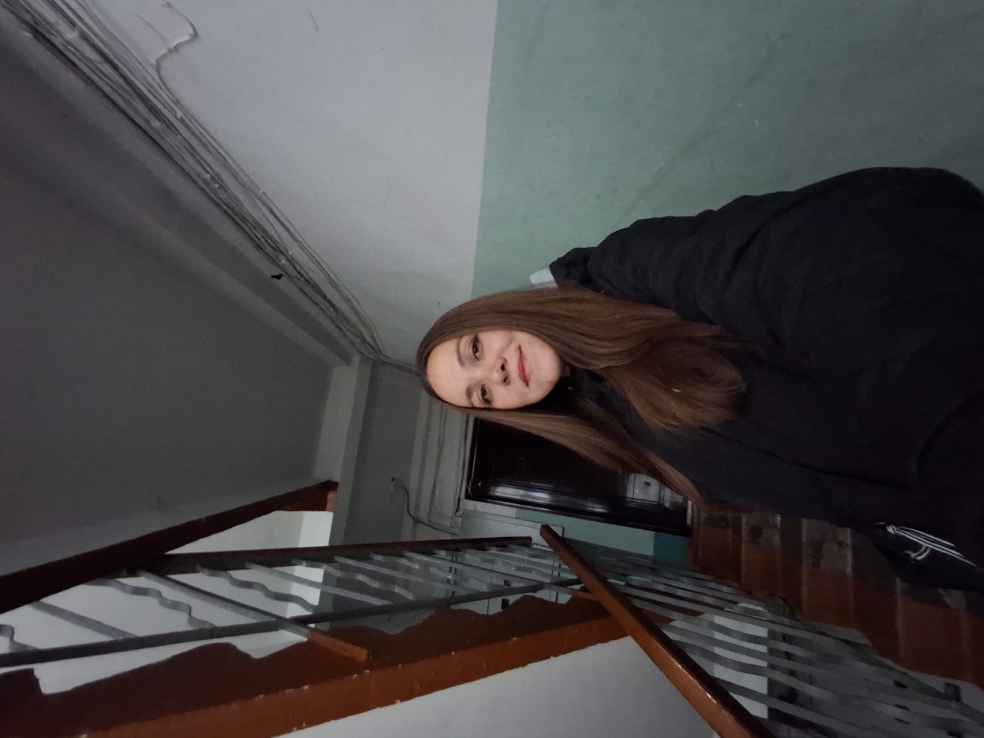An adult female, with long brown hair and a slight smile, is depicted in a low-angle self-portrait within an interior stairwell or hallway in the city of Chaikovsky, Russia. She wears a black padded jacket and looks directly at the camera. To her left, a wooden staircase with worn, dark brown steps and a metal railing featuring vertical bars ascends. The railing is topped by a dark wooden handrail. The surrounding walls are painted in a two-tone scheme: a faded teal/light green on the lower section and off-white/light gray on the upper section, displaying signs of scuffing and wear. Numerous bundled electrical wires are visible running along the wall and ceiling junction in the upper right quadrant of the image. A dark wooden door is partially visible in the background, to the right of the stairs. No other individuals or explicit interactions are visible. The environment appears utilitarian and aged.