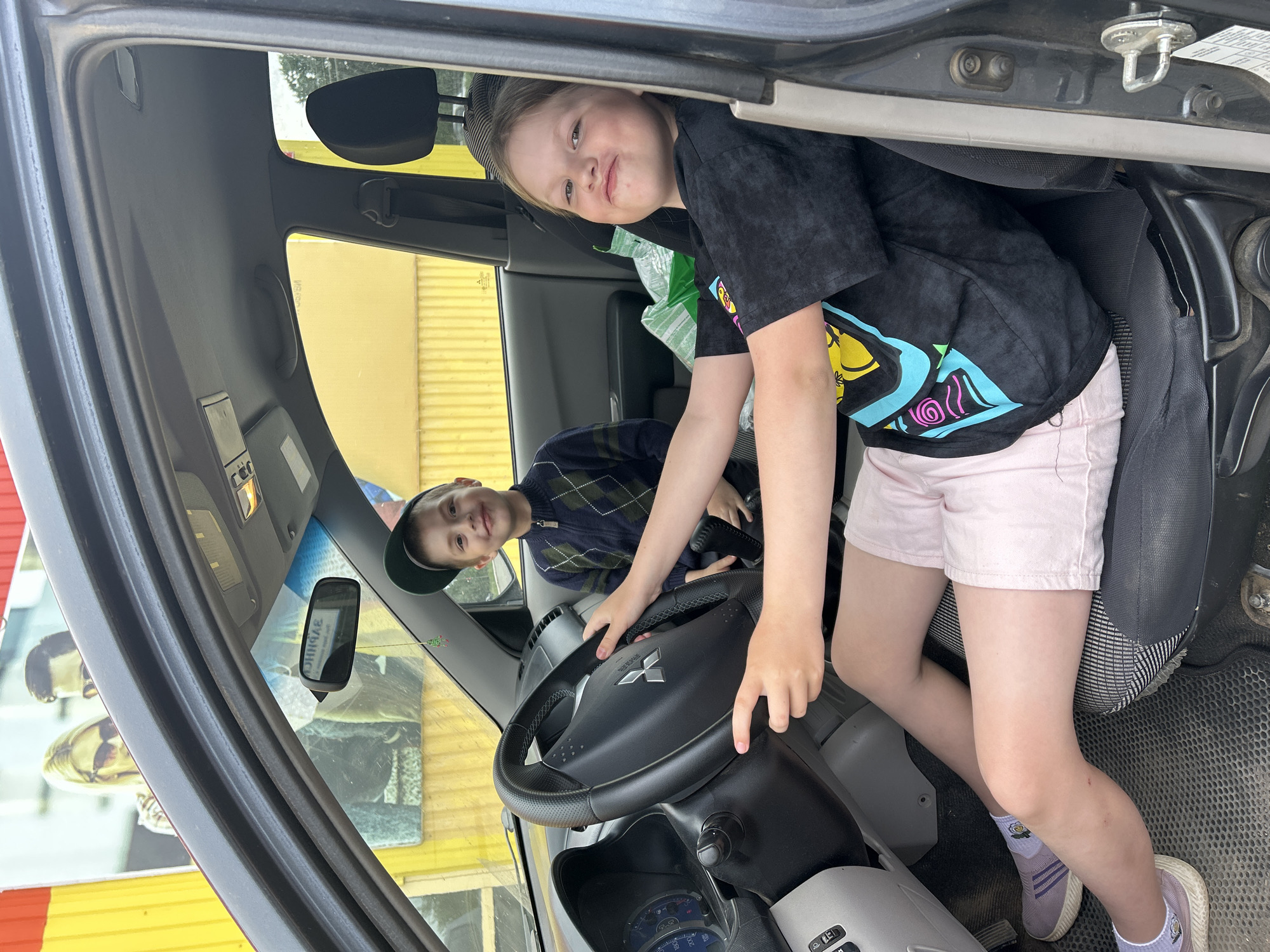 Two children are inside the driver's seat of a parked Mitsubishi truck. A girl wearing a black t-shirt with a colorful design and pink shorts is seated on the driver's seat, her hands on the steering wheel. She looks directly at the camera. A boy in a dark blue sweater and baseball cap is in the background looking toward the camera. The interior of the truck is visible, including the dashboard, rearview mirror, and seats. Outside the truck, there is a yellow building and a promotional poster with pictures of people on it. The photo was taken in Chaikovsky, Russia.