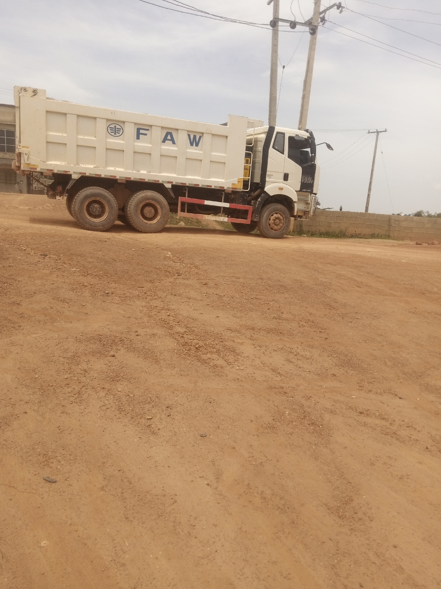 A white dump truck with the FAW logo is parked on a dirt road in Jos, Nigeria. The truck has a large white bed and a black cab. It is a cloudy day, and there are power lines and utility poles in the background. A low brick wall is visible behind the truck, and some sparse vegetation grows beyond it. The ground is covered in reddish-brown dirt and small pebbles.