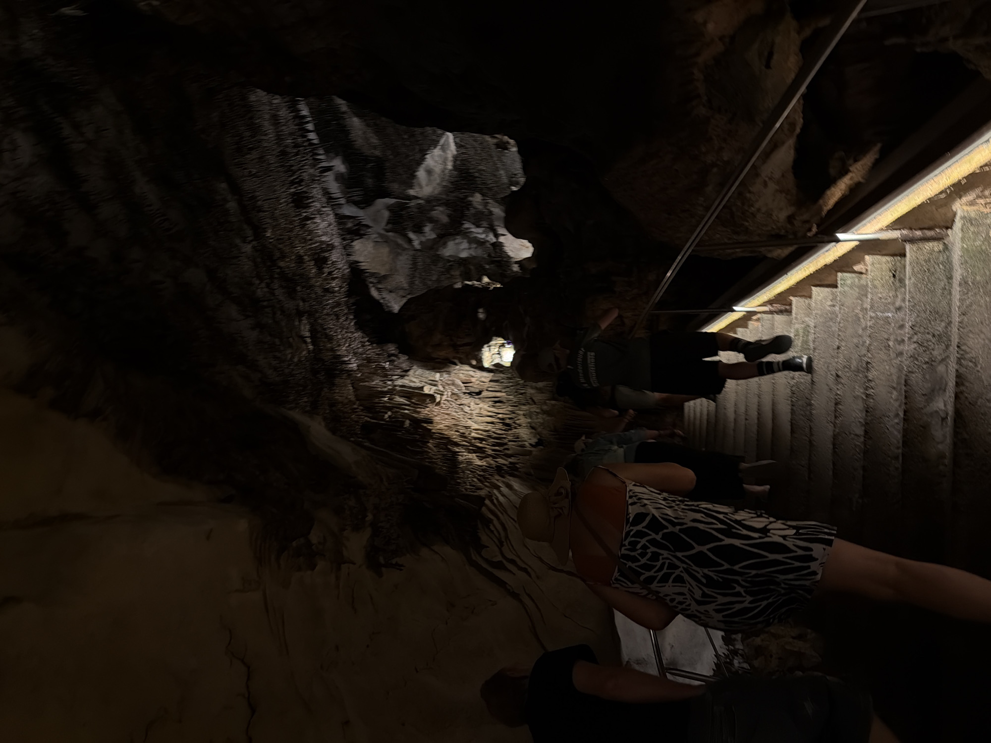 The image depicts a group of people descending a staircase inside a cave, possibly located in Manacor, Spain. The cave features rugged, textured rock formations on the walls and ceiling, illuminated by artificial lighting. A metal railing runs alongside the staircase for support. The people are dressed in casual clothing, with one person wearing a dress with a distinctive black and white pattern. The overall ambiance is dark and cavernous.