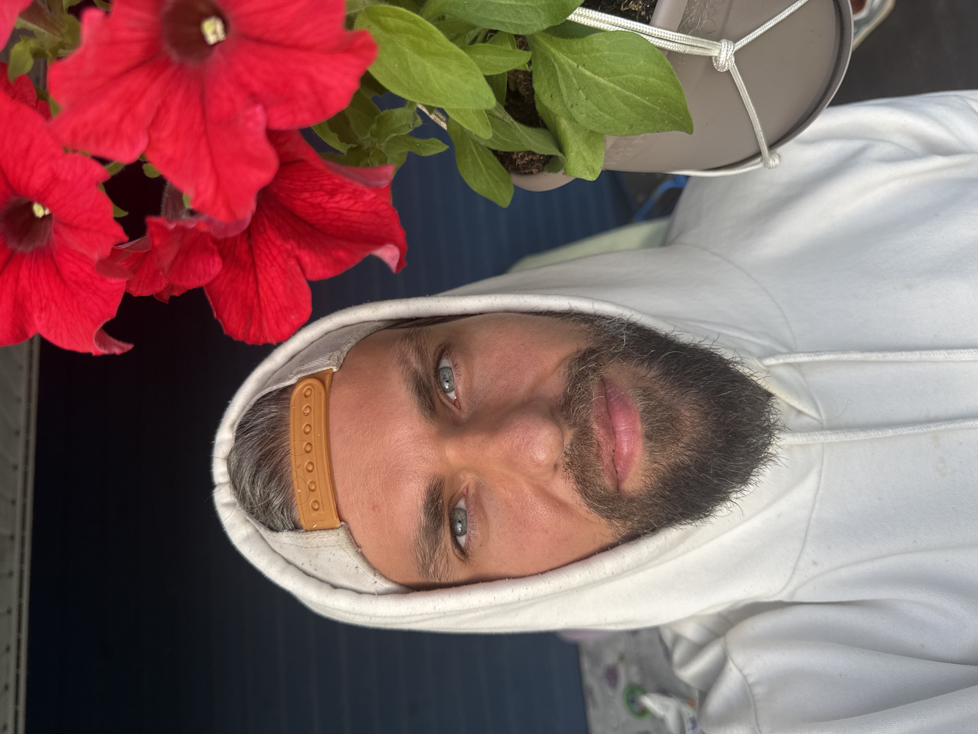 A close-up selfie shows a man with grey-streaked hair and a beard. He is wearing a white hooded sweatshirt and a light-colored baseball cap with an orange adjustable strap. The man has light blue eyes and is looking directly at the camera. To the right of the man's face, a potted plant with bright red petunia flowers is visible. The background is a dark blue surface. The image was taken outdoors, possibly on a porch or patio, given the presence of the plant and the railing visible in the upper left corner. There is no indication that this image was taken in Kuti, Russia.