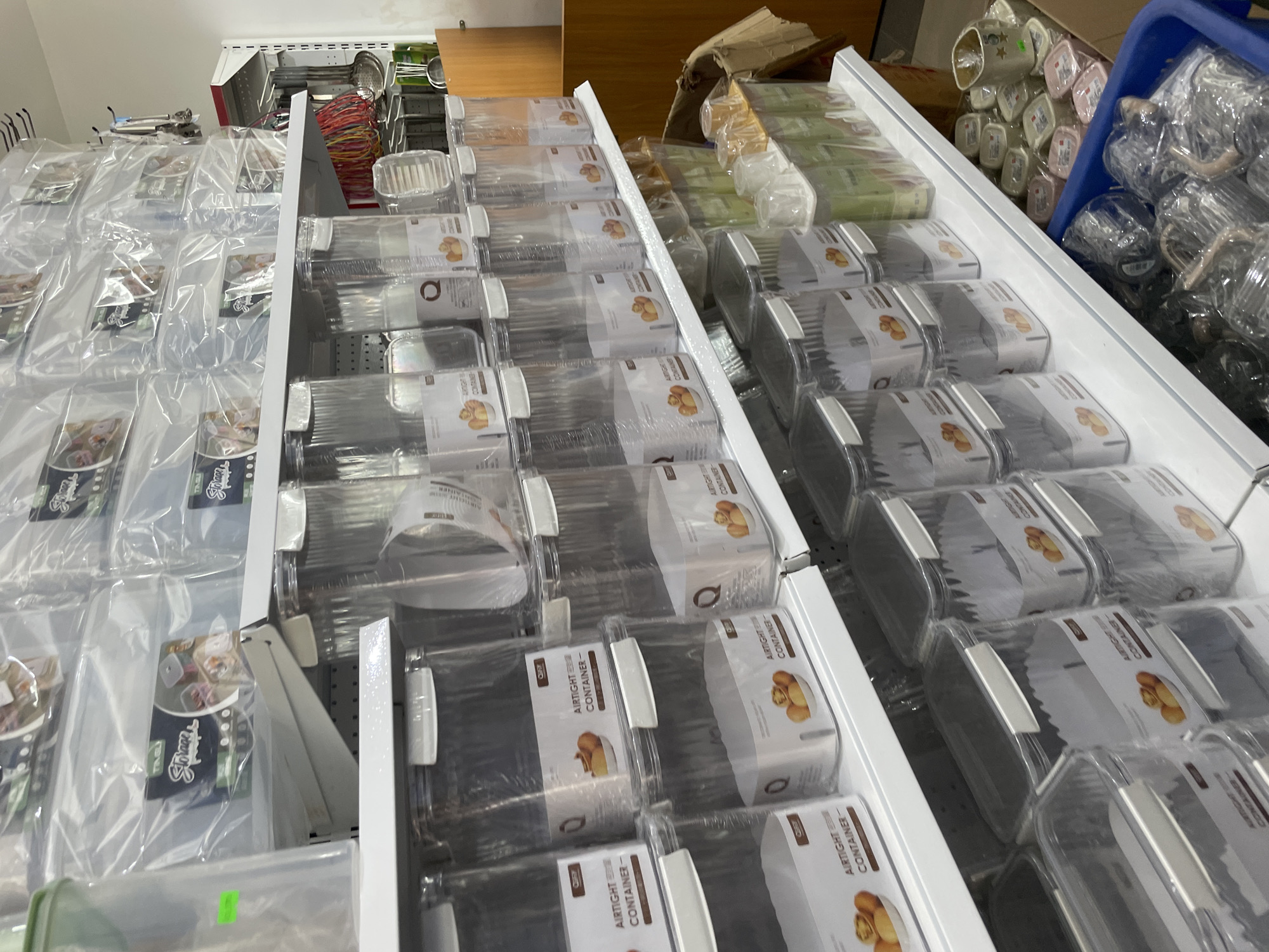 This image shows a display of various food storage containers. On the left are sealed plastic bags containing food products, and on the right are stacks of clear plastic food containers with lids. These containers have labels marked "AIRTIGHT CONTAINER" and include an image of walnuts. In the background, there are other household items and containers. The location cannot be ascertained as Kurunduma, Nigeria from the image itself.