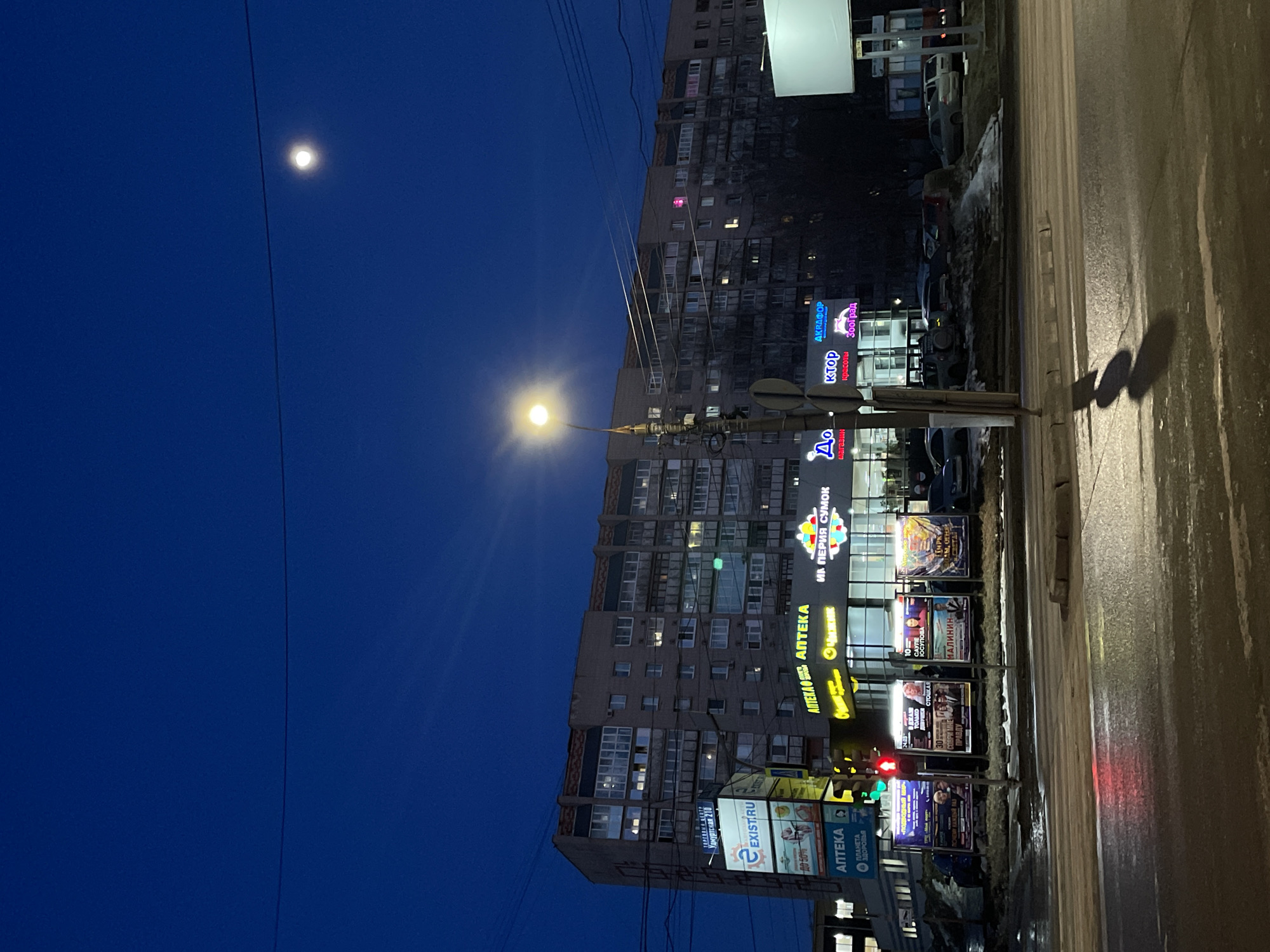 The image depicts a nighttime street scene in Izhevsk, Russia. A tall apartment building dominates the background, with numerous lighted windows visible. Various businesses occupy the ground floor, displaying illuminated signs in Russian. Prominent signs read "APTEKA" (pharmacy), "EXISTIRU," "ИМ ПЕРИЯ СУМОК" (Empire of Bags), "AKRAPOP," and "ктор красоты Зооград" (Beauty Zone). Street advertisements are also visible. A streetlight shines brightly, casting a yellow glow. The sky is a deep blue, with the moon visible in the upper left. A wet, reflective road occupies the foreground, suggesting recent rain or snow. No people are readily discernible in the image.