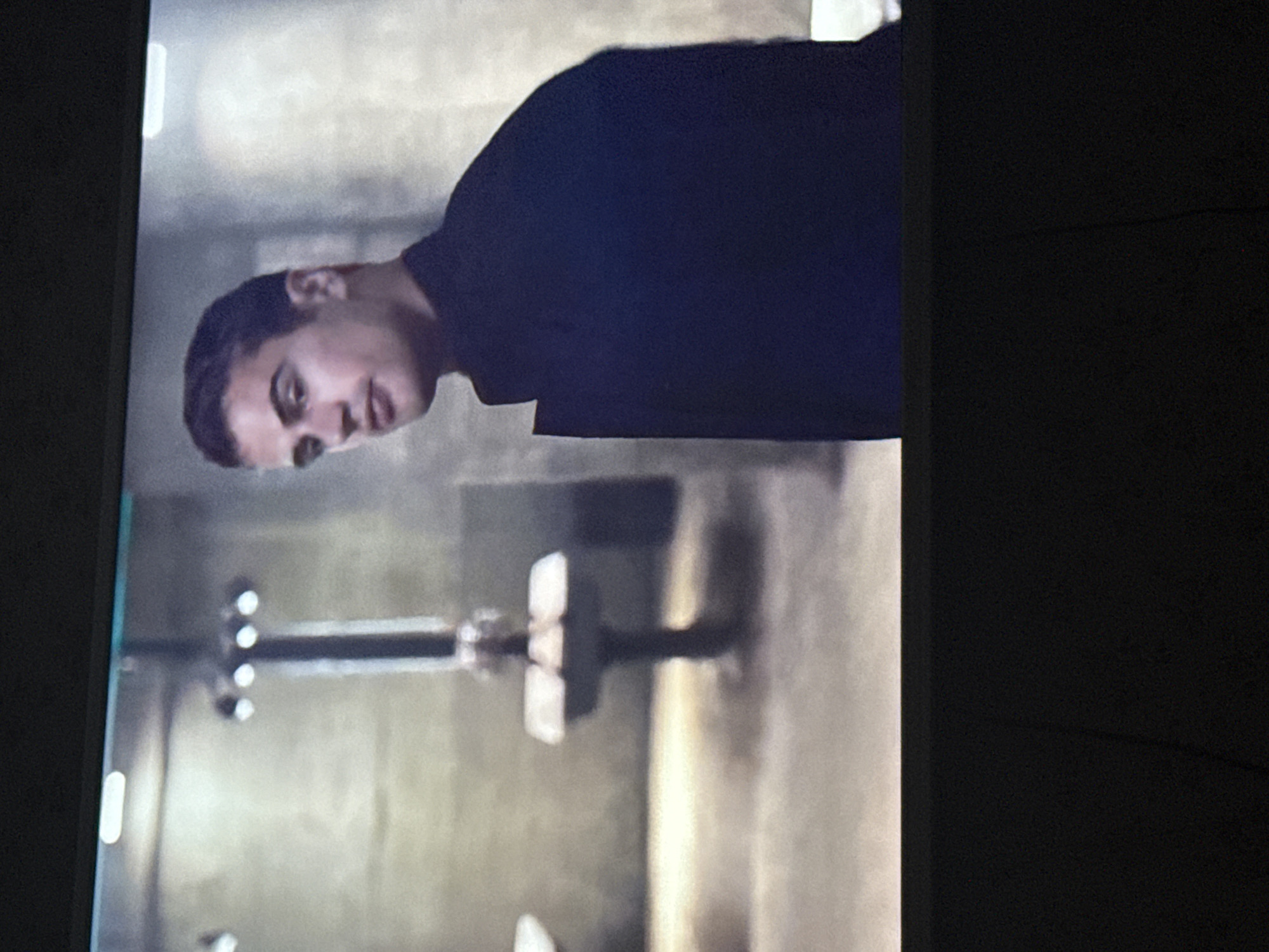 A man in a dark shirt is seen in front of a stainless steel sink and wall with fixtures in what appears to be a bathroom or restroom. There is no indication of the location being in Mytishchi, Russia.
