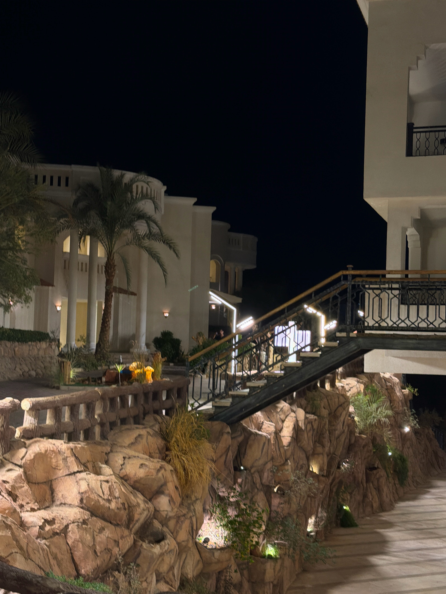 This is a nighttime, eye-level, medium shot of a resort or hotel area in Sharm El Sheikh, Egypt. The scene is dominated by artificial lighting illuminating the architecture and landscaping. On the right side, a building with a white facade and decorative railings on an upper balcony is visible. A dark metal staircase with illuminated steps leads upwards, passing by a rocky, textured wall. The wall is adorned with various types of foliage, including palm trees and dry grasses, with small lights embedded within the rock formations. In the background, a larger white building with classical columns and arched windows is visible, with palm trees in front of it. The sky is dark, indicating it is nighttime. The overall impression is one of a well-maintained outdoor area, likely part of a tourist establishment.