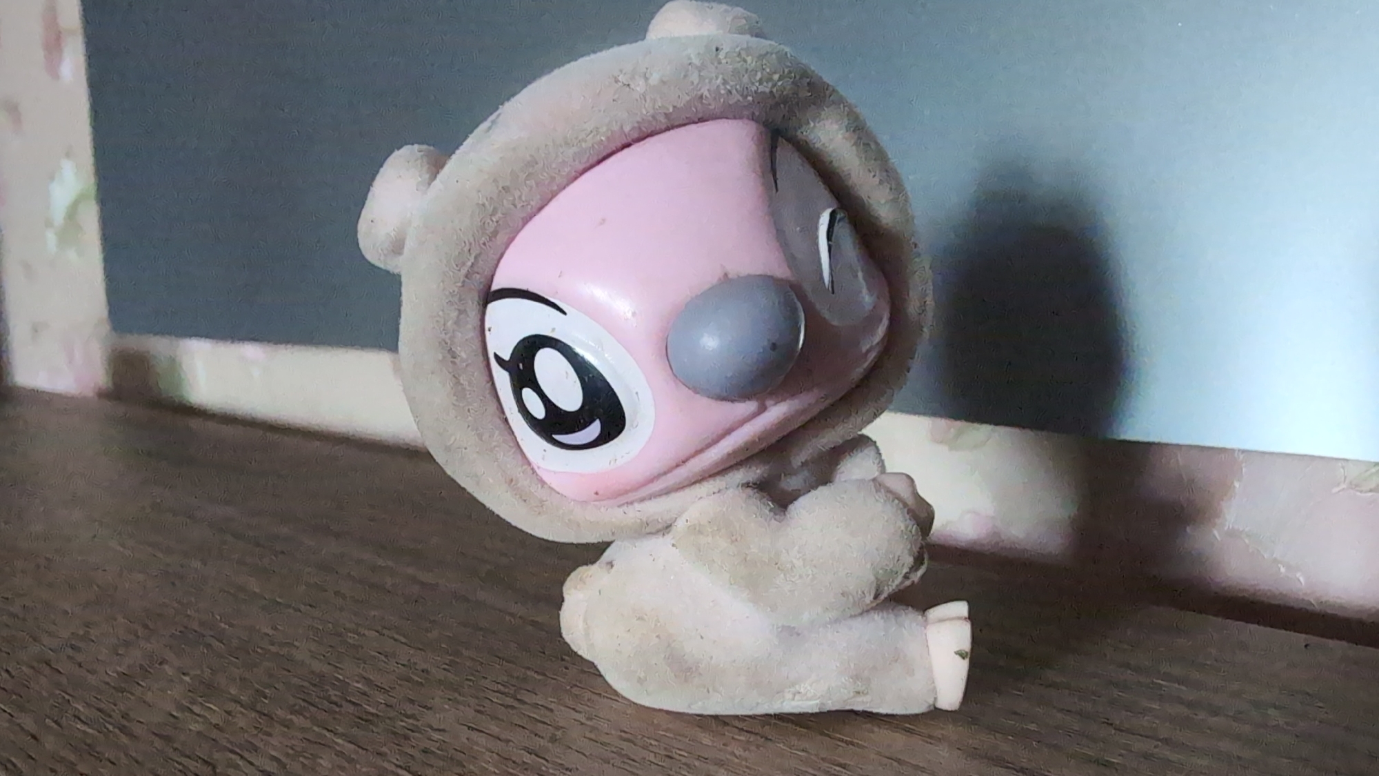 This is a close-up, eye-level photograph of a light brown, fuzzy toy sitting on a wooden surface. The toy has a pink face with large, cartoonish black and white eyes and a gray nose. It appears to be wearing a matching fuzzy hood and pajamas. The toy is angled towards the left of the frame, with its arms clasped in front of its body. Its legs are bent, and it is sitting with its feet pointing to the right.
The background consists of a textured, light blue wall with a narrow horizontal band of pale pink and white patterned wallpaper across the bottom. A shadow of the toy is cast on the wall behind it. The lighting suggests it is daytime, possibly indoors with artificial light, given the sharp shadow.
There is no visible text in the image. The setting appears to be a domestic environment, perhaps a shelf or a table, given the wooden surface and the background details. The toy itself is the sole subject, and the focus is on its details and texture.