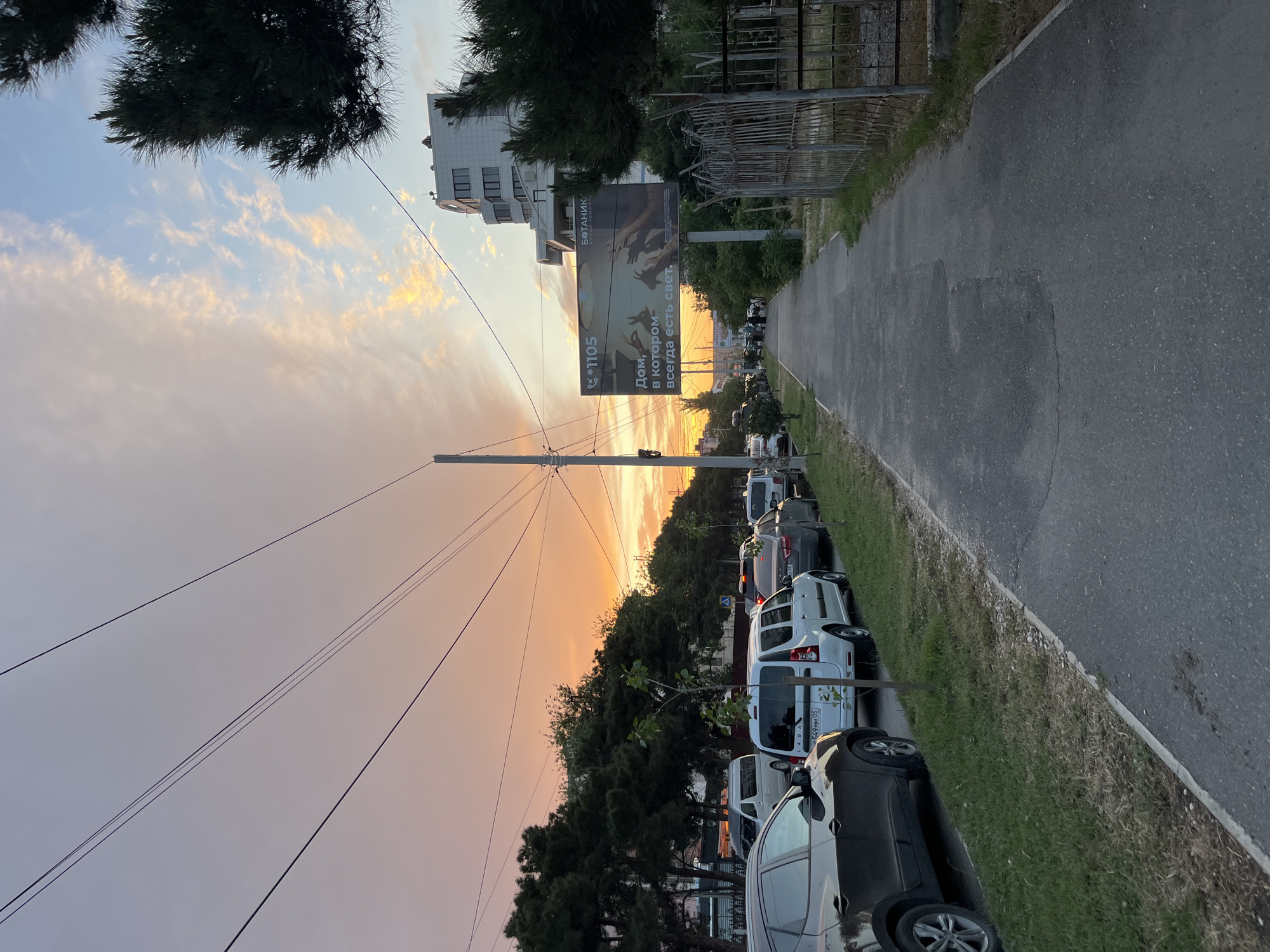 A street scene in Kaspiysk, Russia, at sunset. The image is oriented with the sky to the left. A row of parked cars lines the right side of the road, adjacent to a sidewalk. A tall billboard is visible further down the street. Trees line the road, obscuring some buildings. Power lines stretch across the sky. The sky is filled with clouds tinted orange and yellow by the setting sun.
