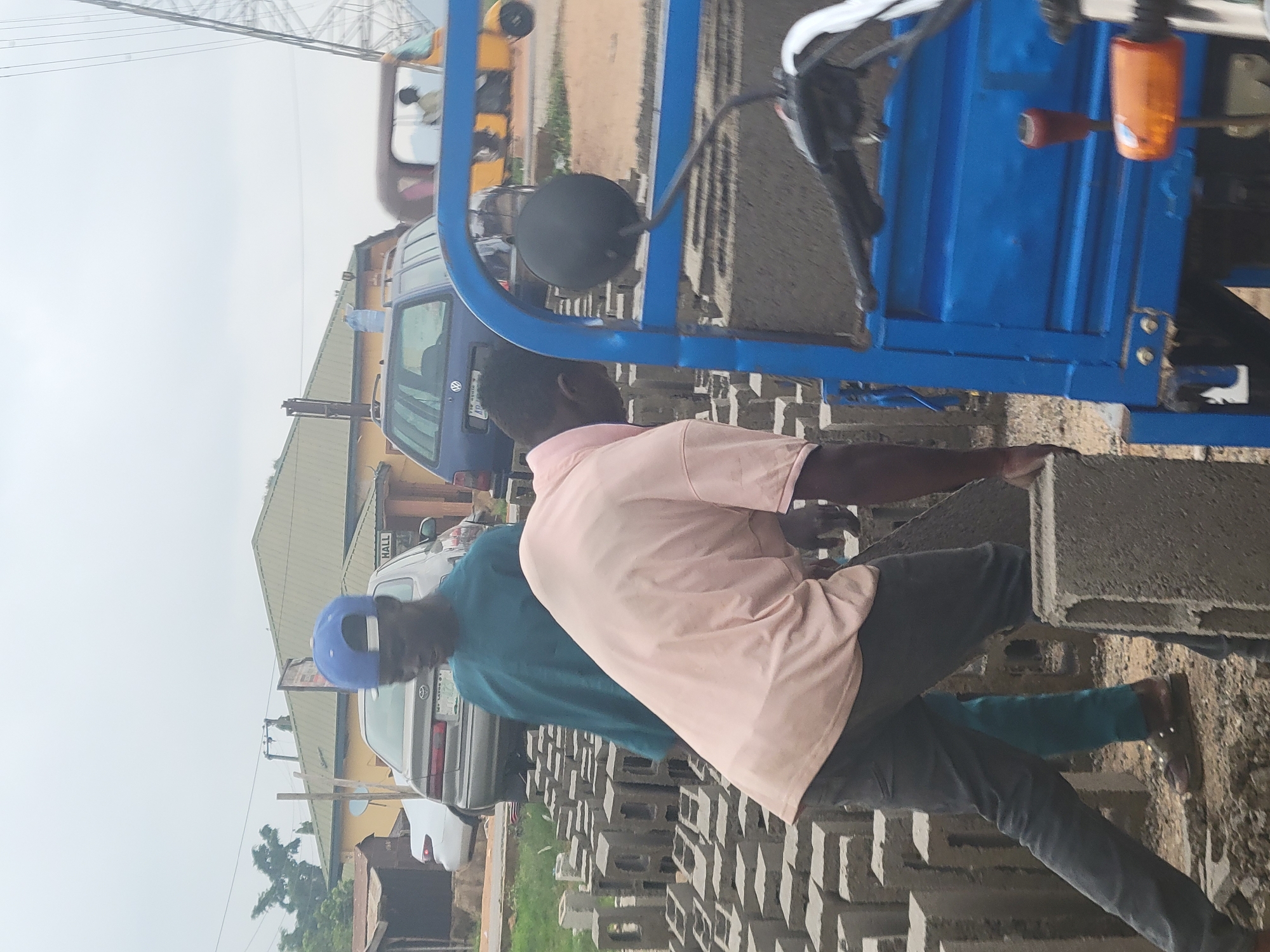 This outdoor scene captures two men engaged in manual labor involving concrete blocks. The primary subject in the foreground wears a light pink short-sleeved shirt and dark trousers, leaning over and appearing to handle a concrete block. Behind him, another man in a teal-colored short-sleeved shirt and a blue cap is also interacting with the blocks, which are stacked in multiple rows on the ground.
In the midground, a blue tricycle, possibly a cargo vehicle, is prominently featured, suggesting it is used for transporting the blocks. A black spherical object, likely a mirror or a light, is attached to the side of the tricycle.
The background shows a street environment with several vehicles. A blue car with a white license plate displaying "LAGOS STATE" and a faint "VW" (Volkswagen) logo is visible. Another lighter-colored sedan is parked further back. Buildings with pitched roofs line the street, and one building has the word "HALL" partially visible on its facade. Overhead power lines or cables crisscross against a bright, overcast sky.
The activity suggests the men are either loading, unloading, or stacking the concrete blocks, likely for a construction-related purpose. The time of day is daytime, under a hazy or overcast sky, providing soft, diffused light. The overall setting indicates a functional, possibly commercial or industrial area in Kwamba, Nigeria.