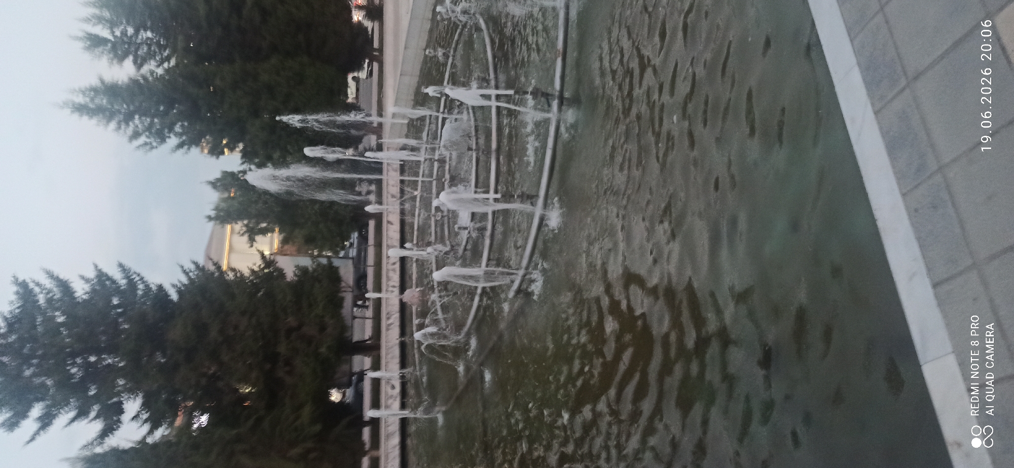 The image shows an outdoor fountain with multiple streams of water spraying upwards in a circular pattern. The fountain is located in what appears to be a public space, with tall evergreen trees surrounding it. In the background, there are buildings and some parked cars, suggesting an urban or suburban setting. The lighting suggests it is either early morning or late evening, as the sky is overcast and the light is soft. The water in the fountain is dark and appears somewhat murky. The foreground shows a paved area next to the fountain, with a patterned surface. A timestamp and camera model are visible in the bottom left and right corners of the image, reading "REDMI NOTE 8 PRO AI QUAD CAMERA" and "19.06.2026 20:06" respectively. There are no people visible in the image.