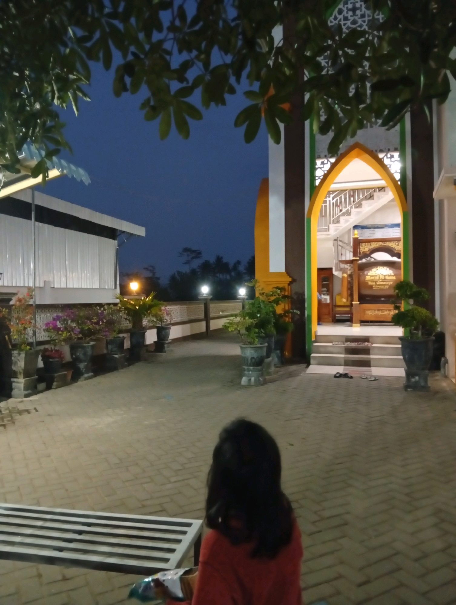 At dusk in Wonocoyo, Indonesia, a young person with long dark hair, wearing a red shirt, sits on a slatted bench facing away from the camera. The bench is made of metal and is positioned on a paved courtyard of tan and grey interlocking bricks. To the right of the bench, a mosque entrance is visible, adorned with an arched doorway featuring orange, brown, and green accents, and ornate white latticework above. Inside the arch, a staircase leads up to a wooden structure with Arabic calligraphy. The courtyard is lined with potted plants and flowers, and illuminated by soft artificial lights. A white building with a yellow overhang is situated to the left of the scene. The sky is a deep blue, indicating twilight.