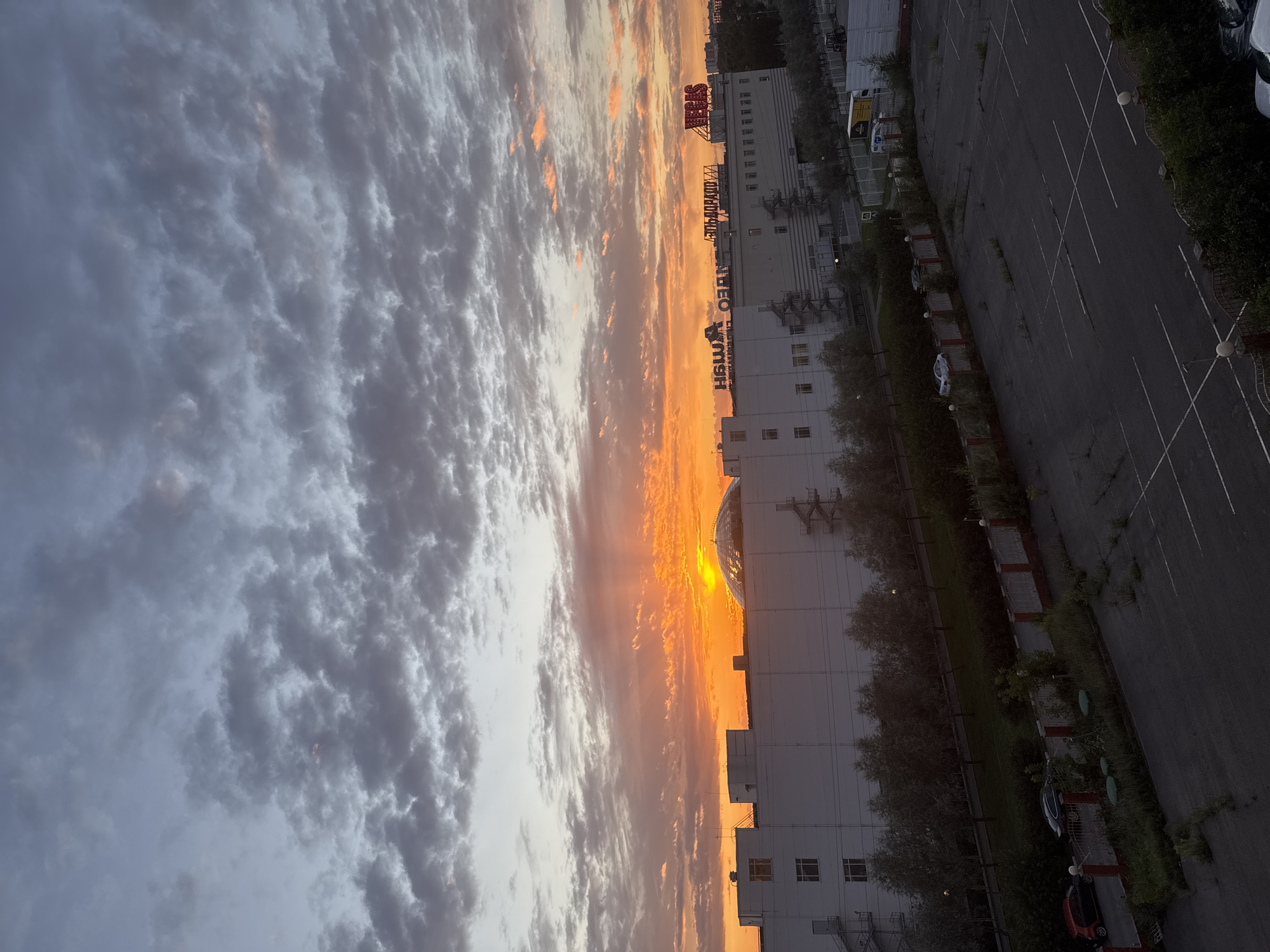 The image captures a sunset over an urban landscape in Sovkhoza imeni Lenina, Russia. The sky is filled with dramatic clouds, ranging from deep grey to vibrant orange and yellow hues, illuminated by the setting sun. Directly below the colorful sky, a large, white, multi-story building dominates the mid-ground. This building appears to be a commercial or industrial structure, featuring multiple windows and external staircases. Several signs are visible on the roof of the building, including one with red lettering that reads "VEGAS". To the left of the building, a large dome structure is partially visible. A row of trees lines the front of the building, and beyond that, a fenced area separates it from a nearly empty parking lot. Several cars are parked in the lot and along a road in front of the building. The foreground shows the surface of the parking lot with faded white lines indicating parking spaces, and overgrown weeds. The overall scene depicts a typical evening in a developed area, with the natural beauty of the sunset contrasting with the man-made structures.